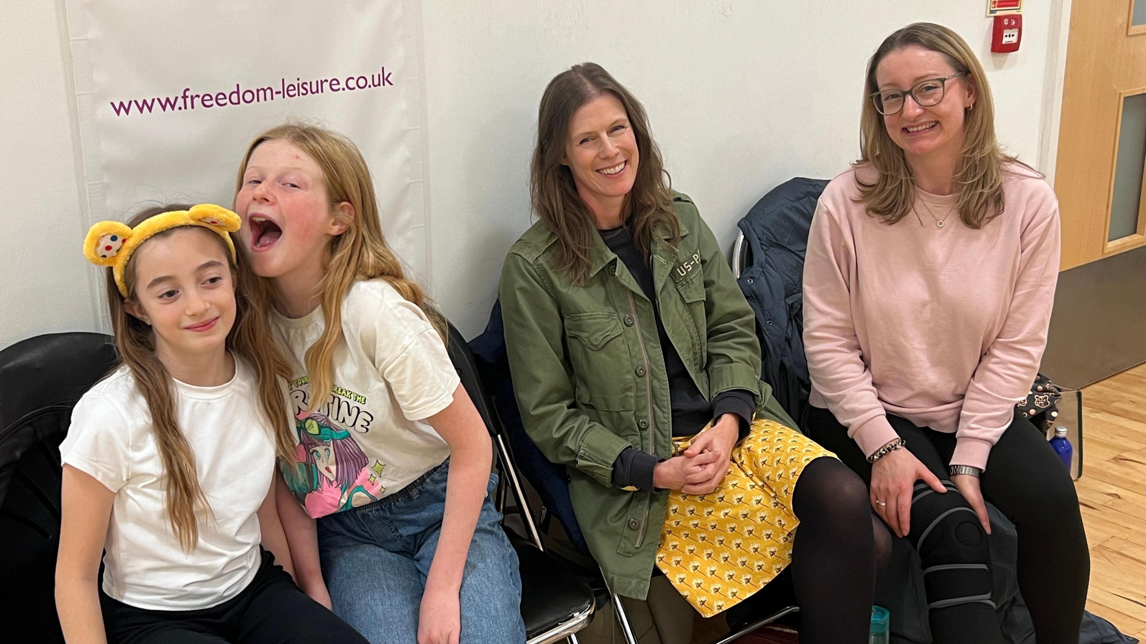 Girl with yellow Pudsey ears sitting with friend pretending to bite one of the ears next to their mums. 