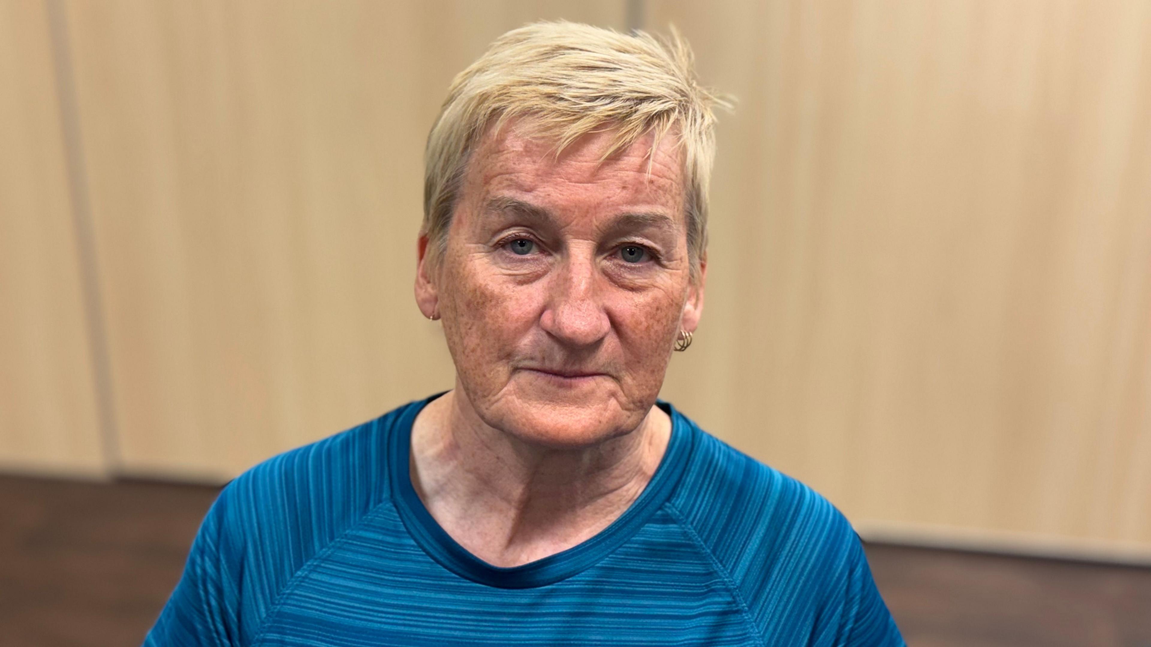 A woman with short blonde hair is looking into the camera. She is wearing a blue striped top and gold hoop earrings.