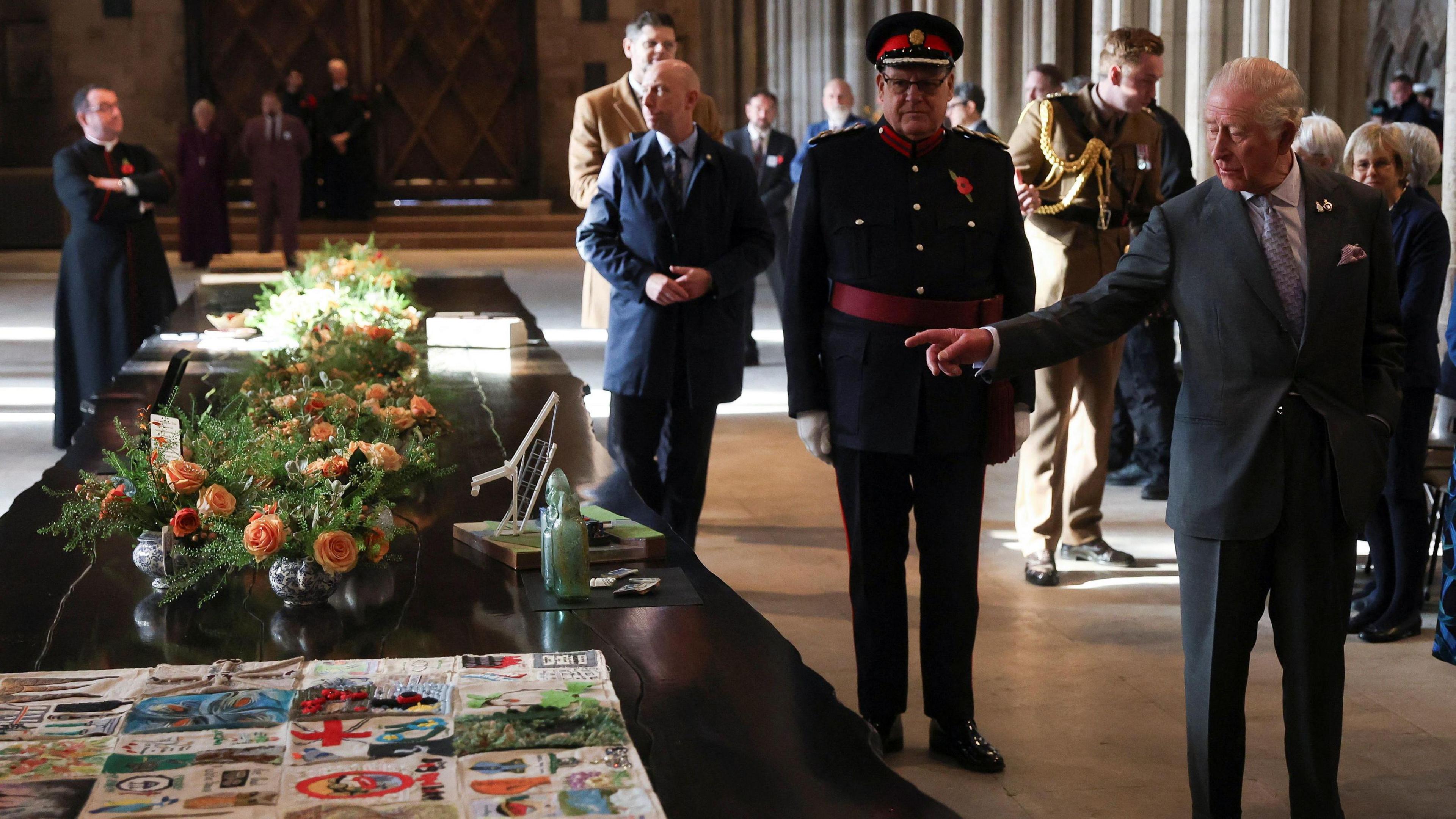 King Charles stands to the right of a long black table, which has flowers and artwork on it. People in suits and military uniforms are stood behind him.