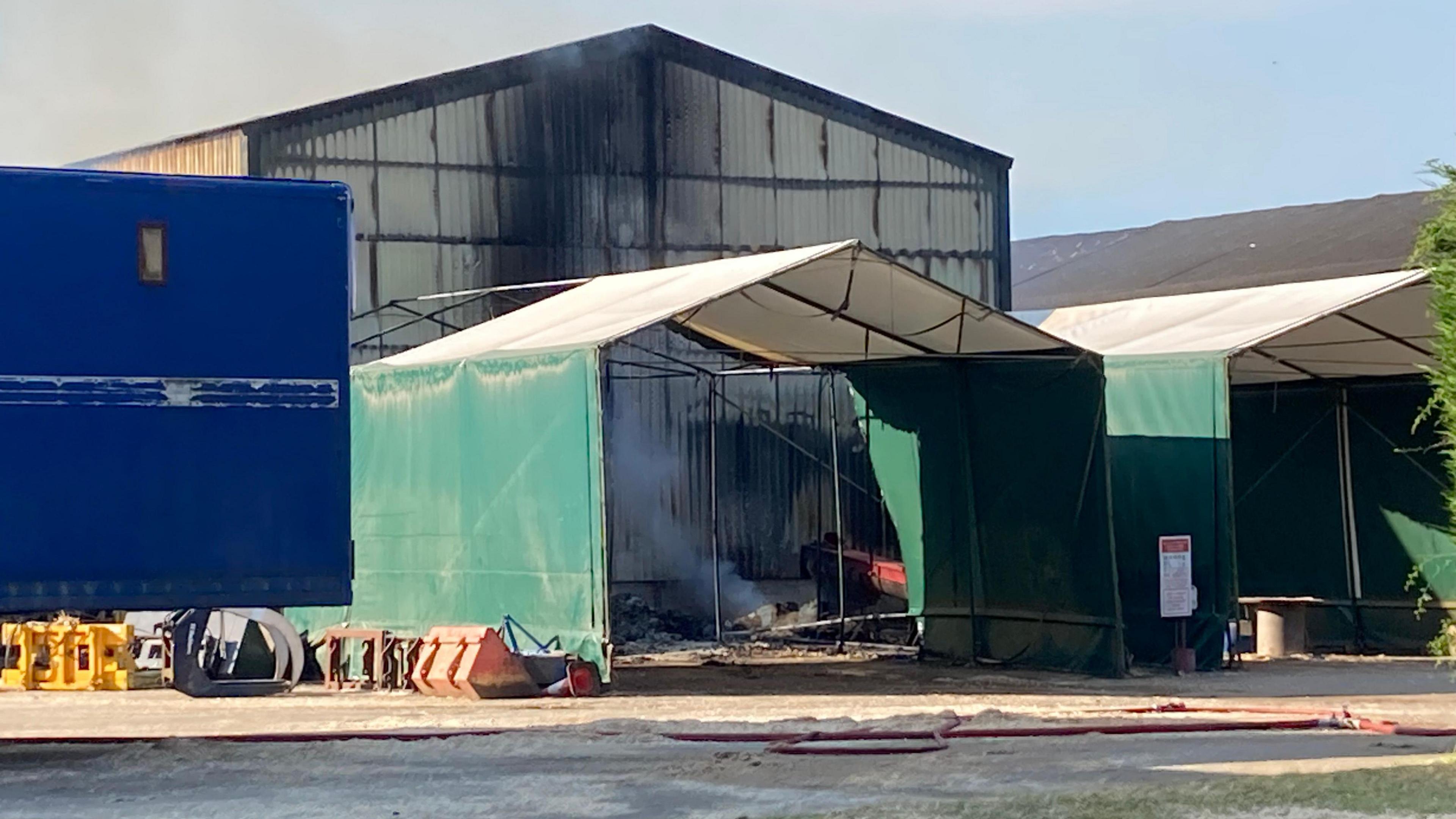 The back of two large corrugated metal barns partly blackened by smoke. Behind them are large open gazebos and the back of a blue lorry or van.