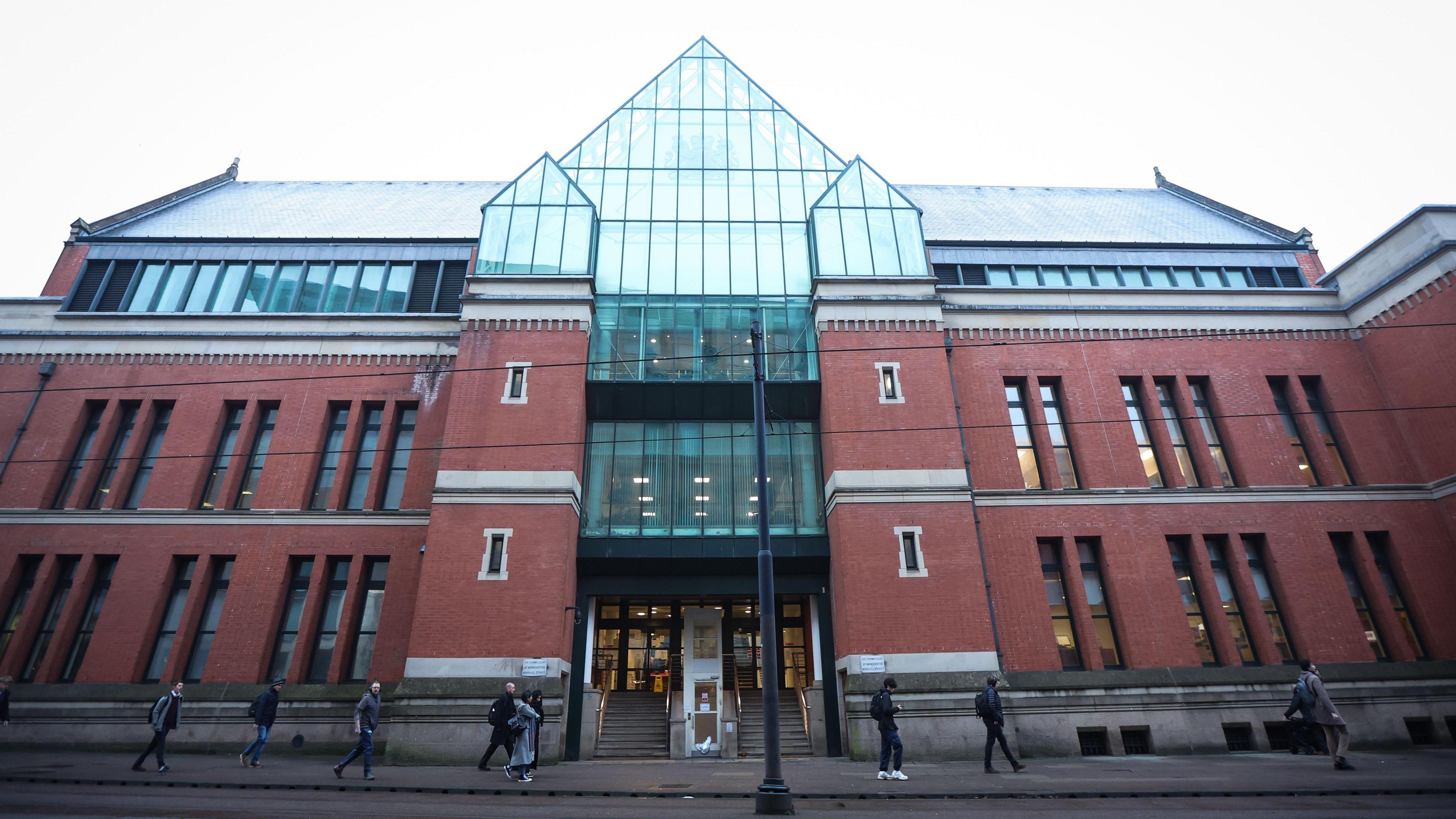 The entrance to Minshull Street Crown Court in Manchester. with two sets of stairs in between two columns of the redbrick, multi-storey building, on top of which sits a wall of glass panels rising to a point. People can be seen walking along the pavement outside. 