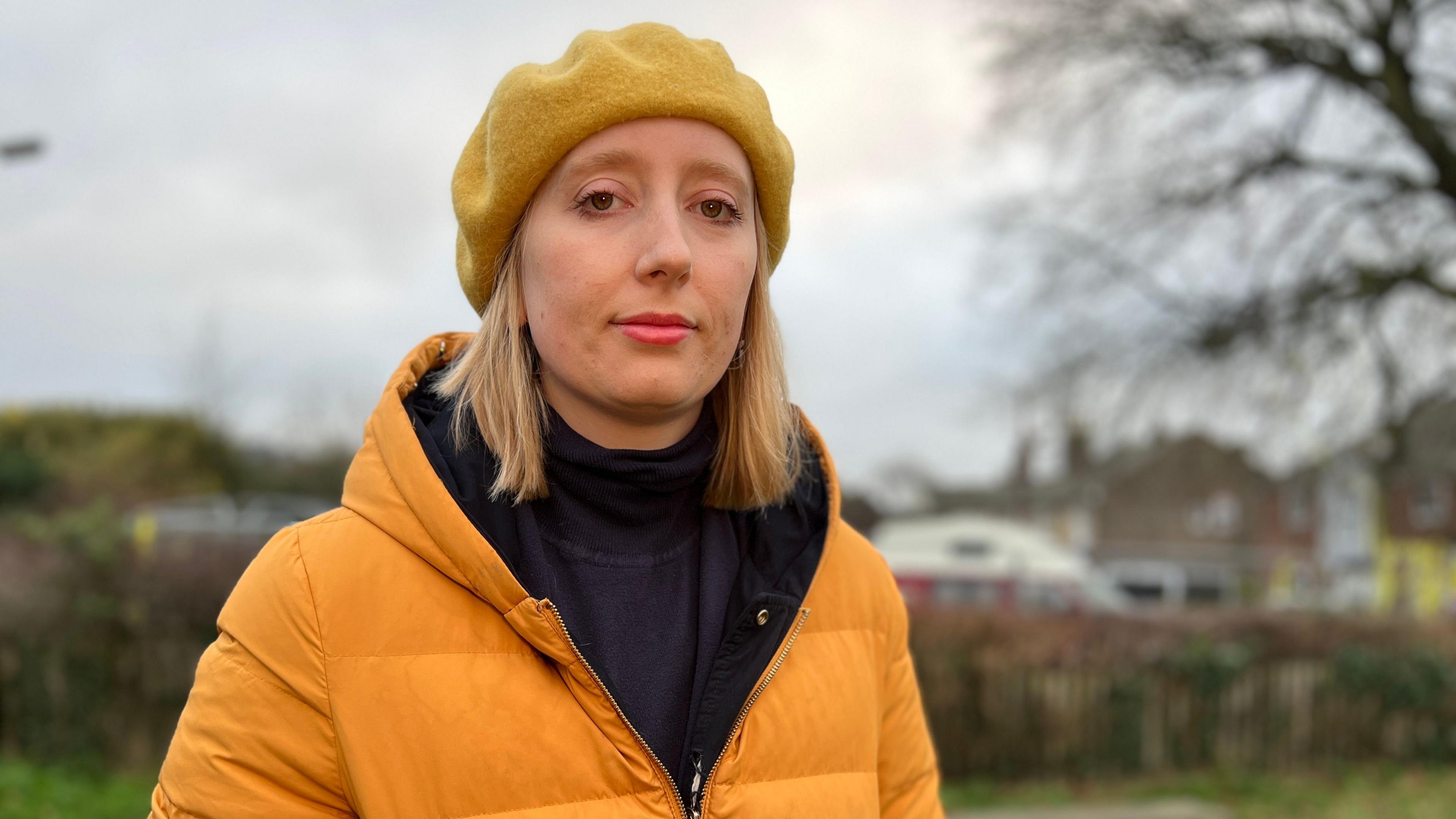 Kat Scott, a Wivenhoe resident, stood in a park with a tree behind her. She is wearing an orange coat and an orange beret.