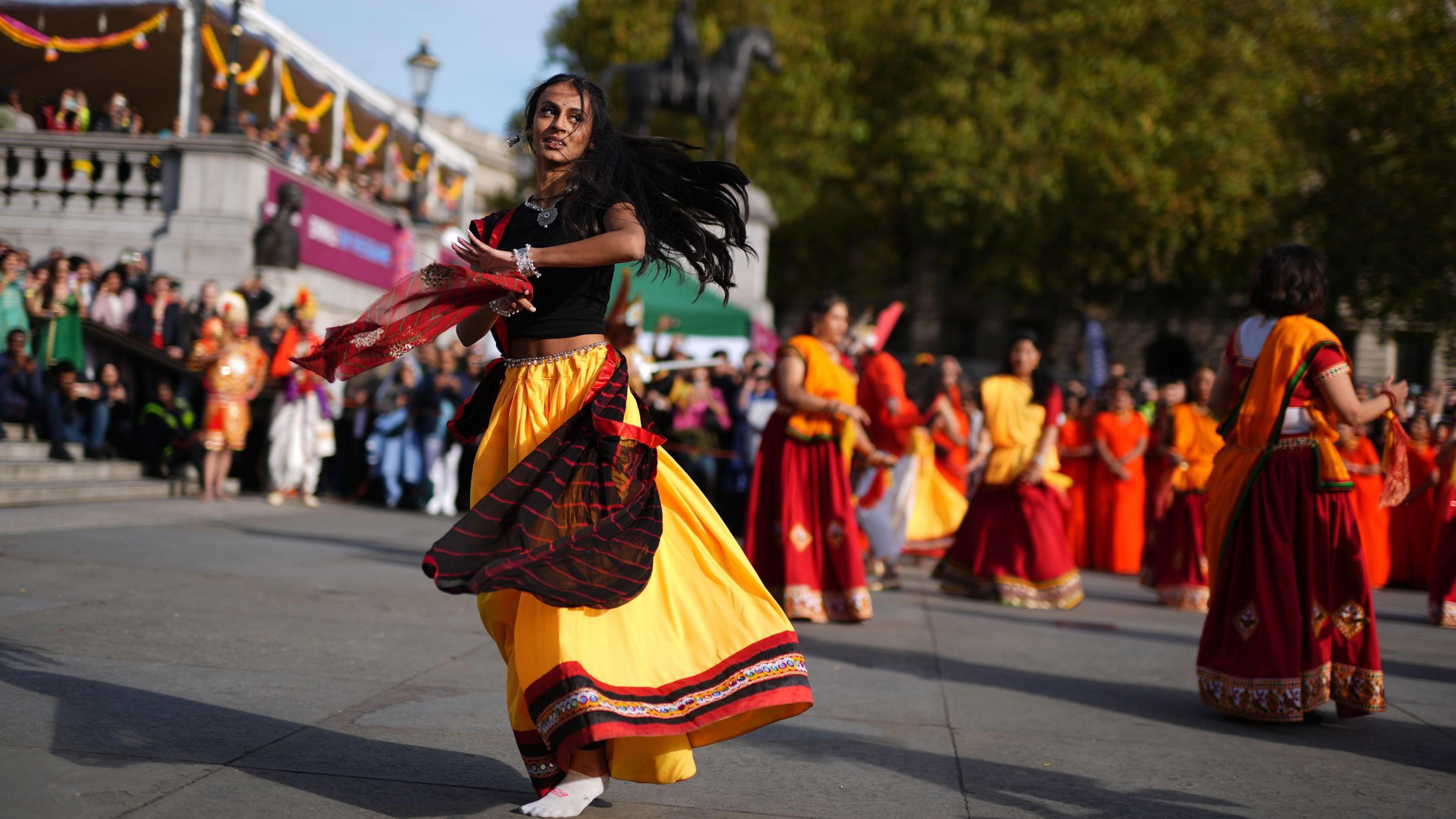 a woman wearing a long skirt and a sari dances in trafalgar square while people watch from afar, other dancers wearing bright colours are in the background