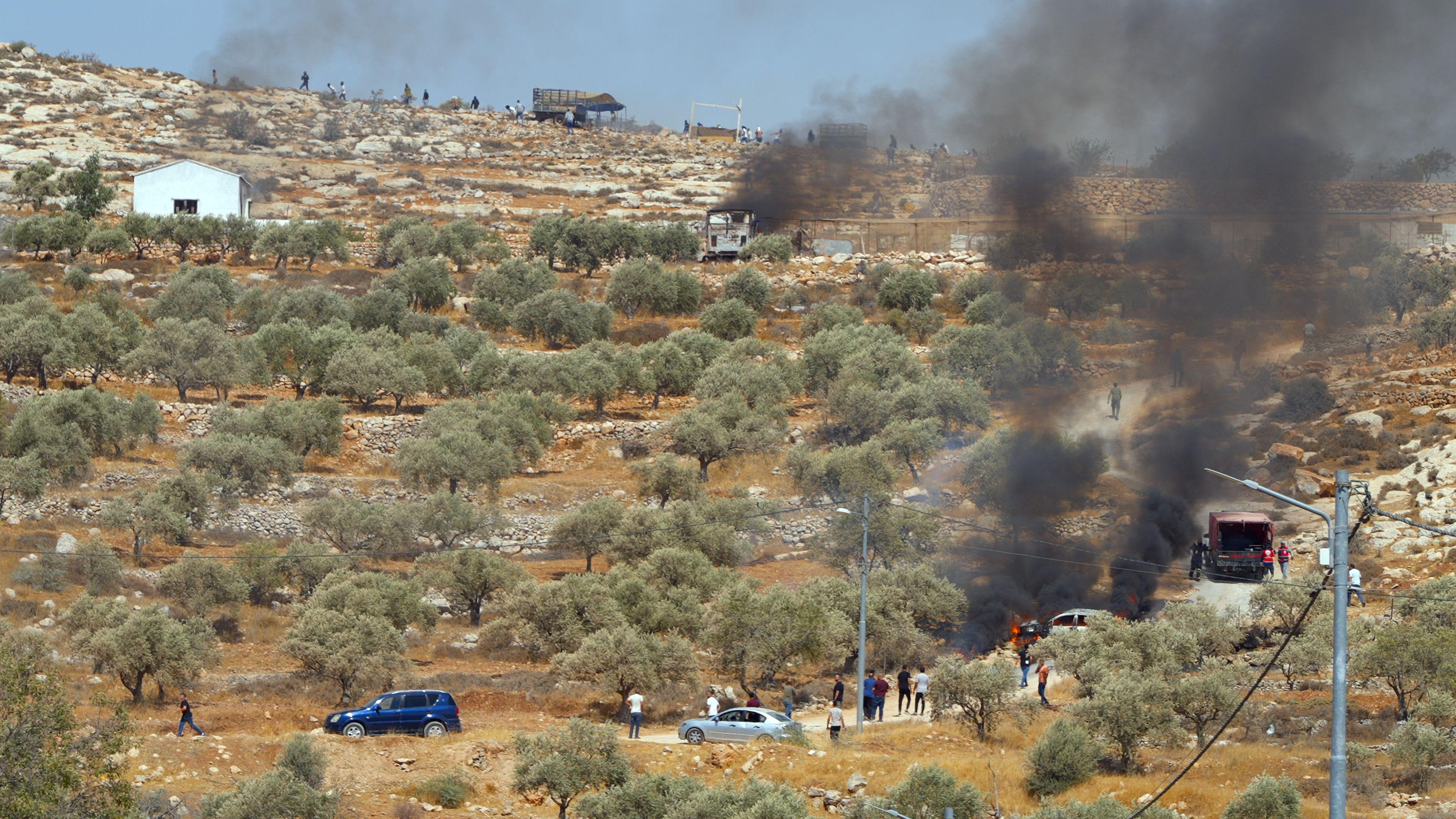A large expanse of land with cars dotted across. Dark smoke billows up towards the sky in the right hand corner where a fire rages on. 