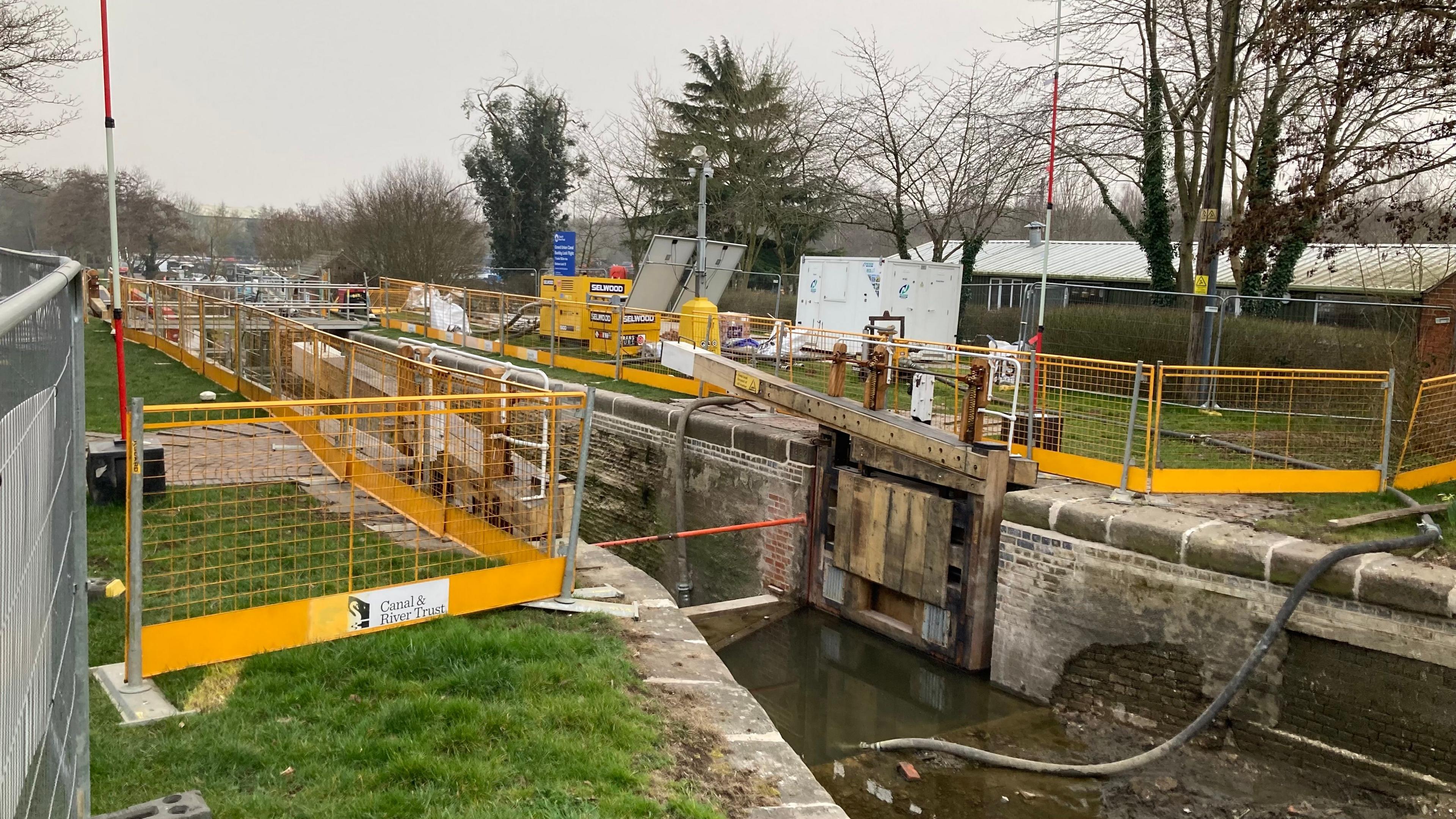 A canal lock. An open gate is visible and the area is surrounded by yellow railings. There is a pipe into the water from the opposite towpath. There is a grass bank on either side of the canal and trees are also visible.