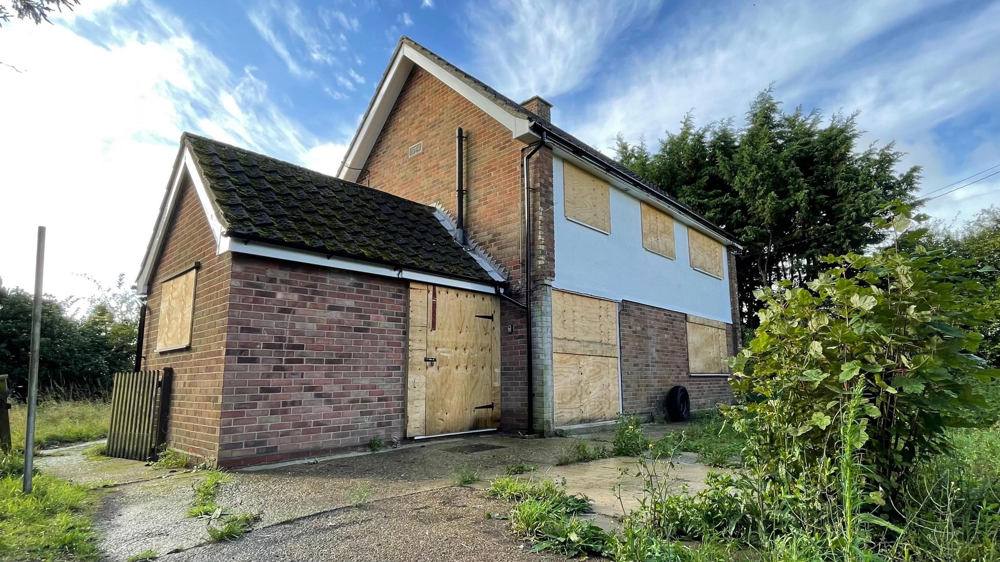 A detached, brick-built house, whose windows and doors have been boarded up. The picture is looking diagonally towards the house, which has three windows on an upper floor. It is the only building visible in the image. A foundation stone, just visible on the gable end, says it was built in 1967. There is concrete hardstanding around the property, which has grass and weeds growing through it in places. A small bush is seen in the right-hand foreground, with trees and lawn in the distance. A blue sky, streaked with some cloud provides the backdrop.