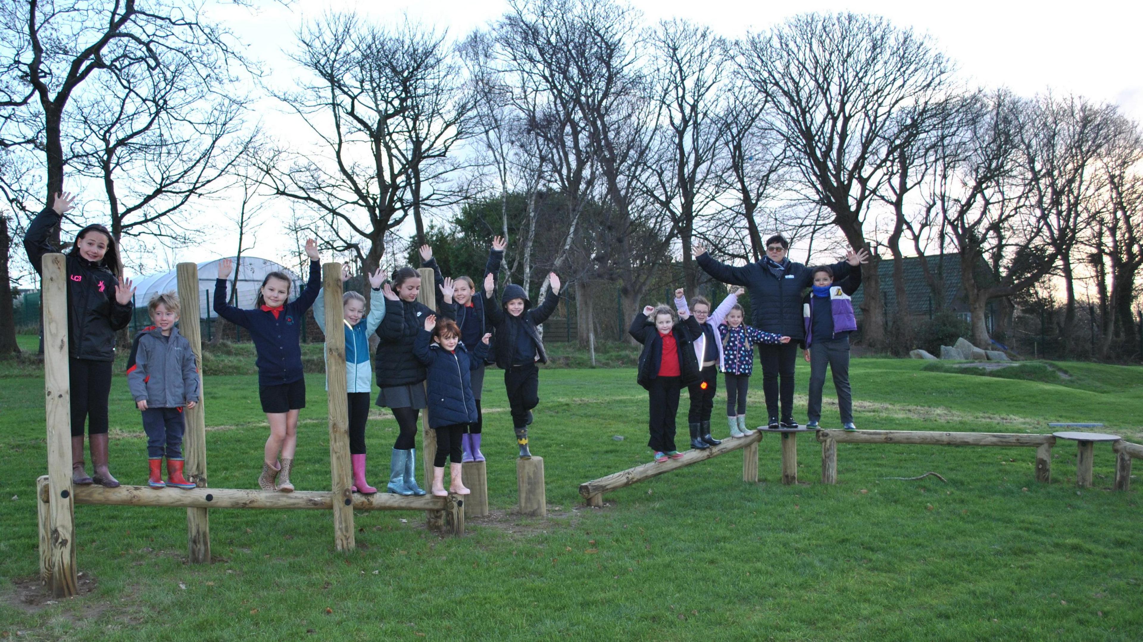 A group of children standing alongside a woman with short brown hair on wooden beams, which make up part of the new adventure trail, in a school field. They are wearing coats and wellies and holding their hands up in the air in the air.