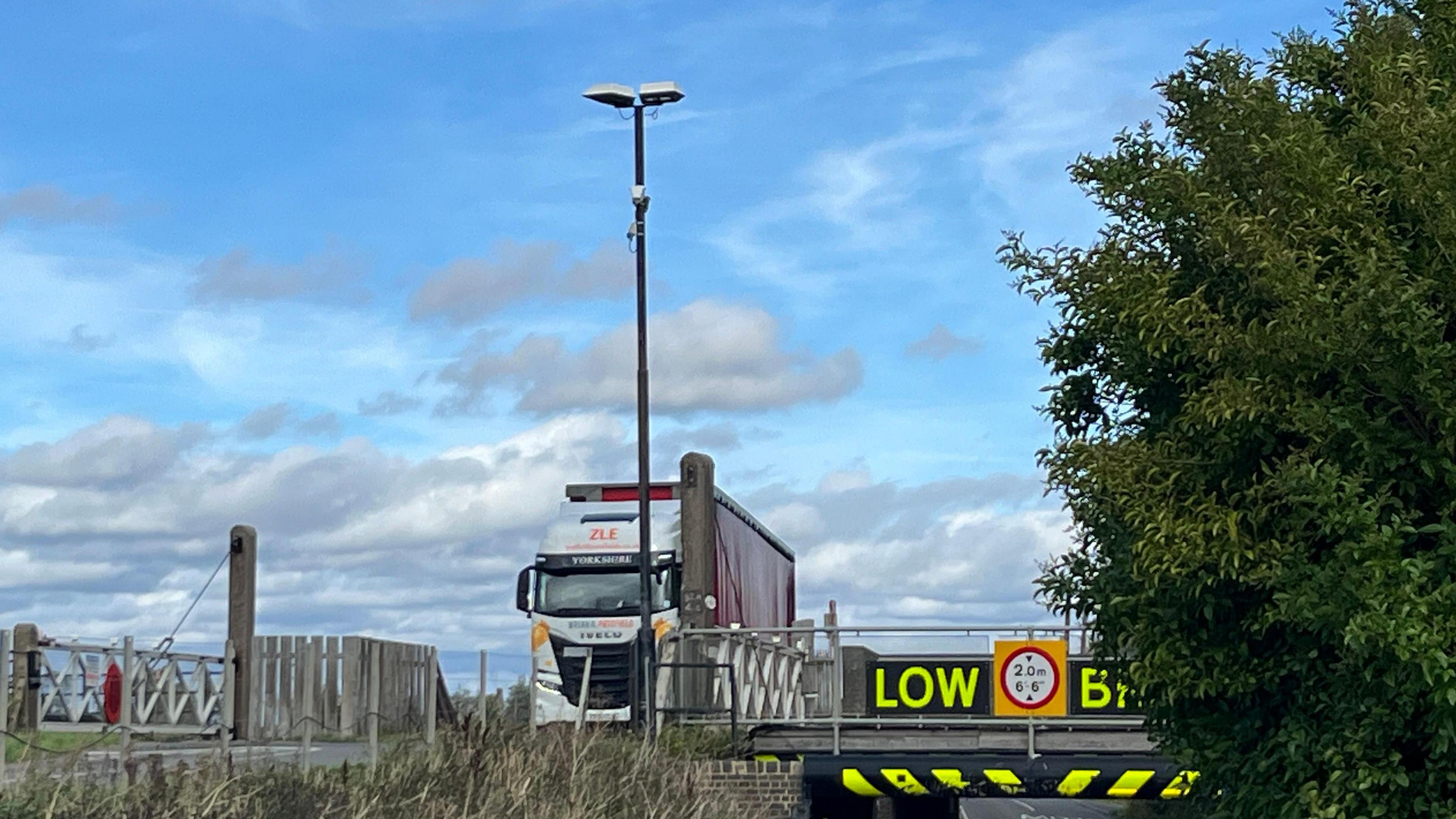 A lorry passing over a level crossing - which is over the bridge and railway. The bridge is lower down to the right of the crossing and has large yellow letters, reading "low bridge". Between the two is a long metal pole with lights on top and a small white camera high up - facing down on to the road and bridge. 