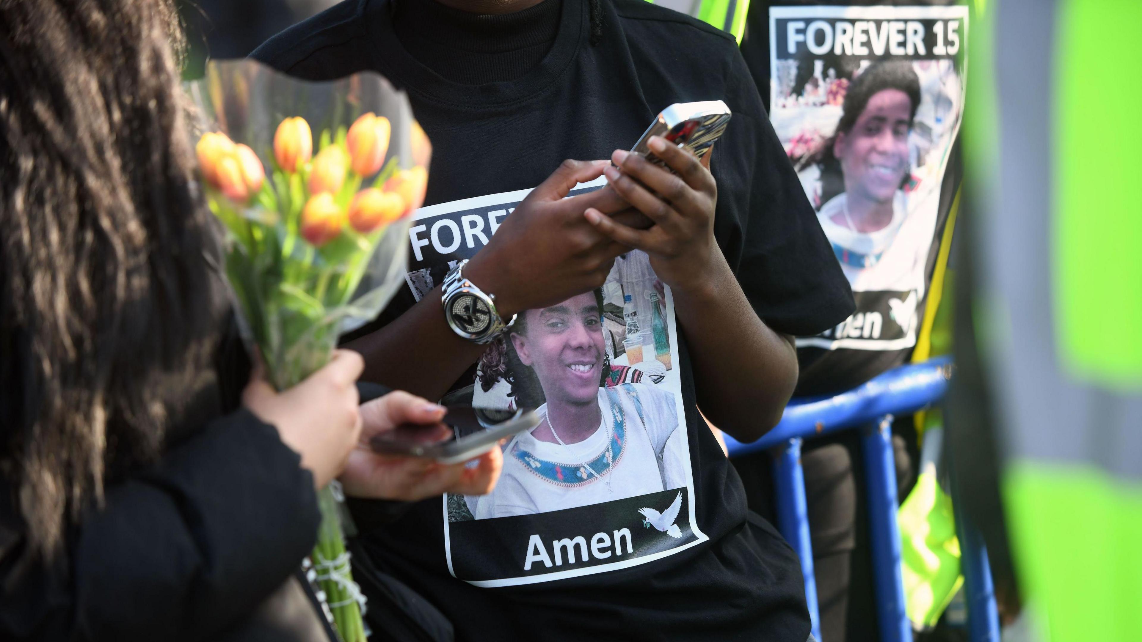 A young man or boy, his head and face out of frame, is doing something with his phone, possibly taking a picture of something on the ground. He is wearing a black t shirt with a photo of Amen and the legend "Forever 15 - Amen" on the front. A person behind him is wearing a similar T-shirt. Another person closer to the camera and slightly out of focus is using their phone and carrying flowers.