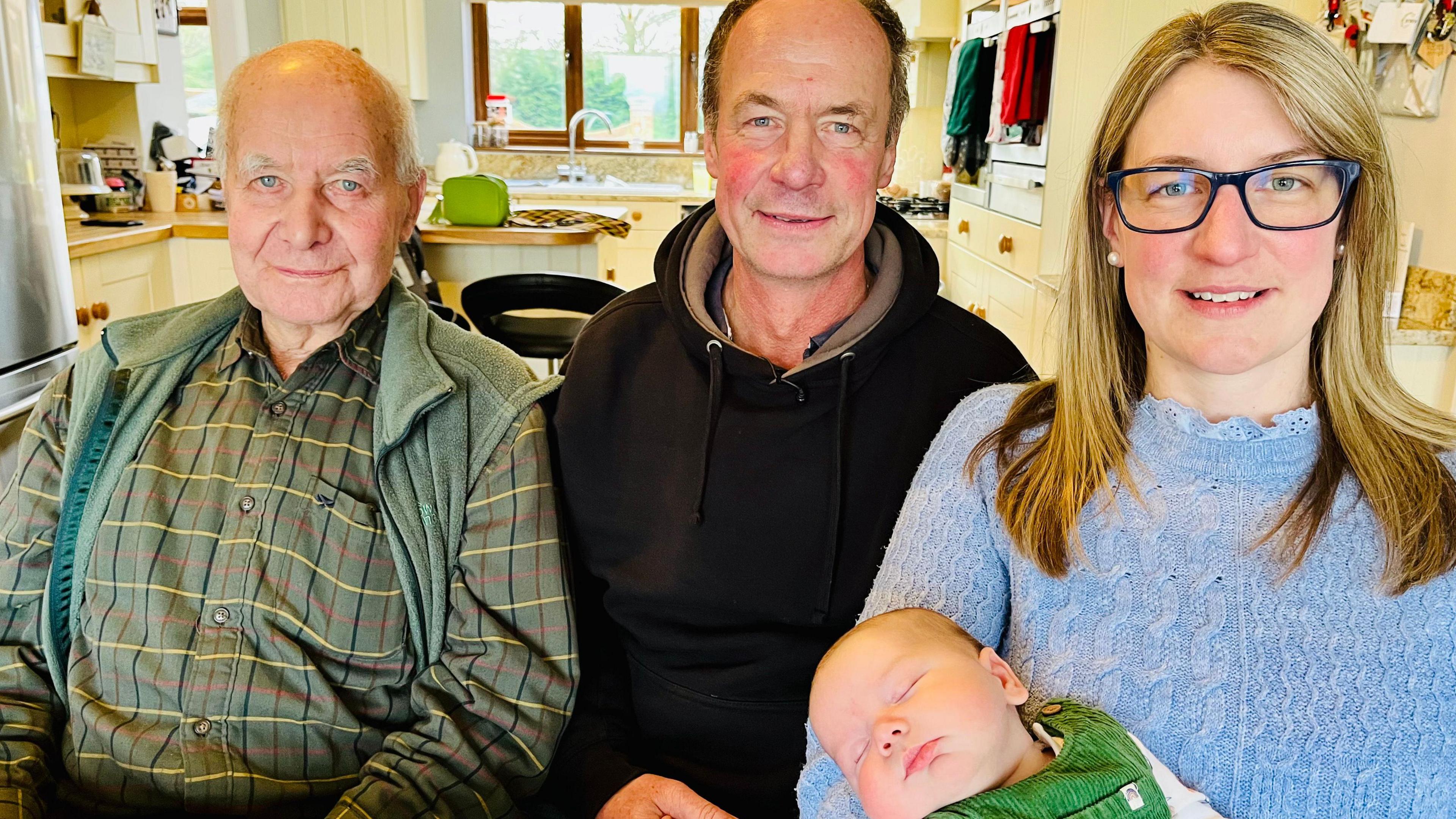 A family of four sitting in a kitchen looking into the camera. Bob has a green shirt with thin yellow, red and blue horizontal lines and a green gilet. He has short white hair and is bald on top. David is wearing a black hooded jumper and has receding dark hair, while Kate has blonde hair to just below the shoulders and is wearing dark blue-rimmed glasses and a light blue jumper. She is holding her baby son, who has a green outfit