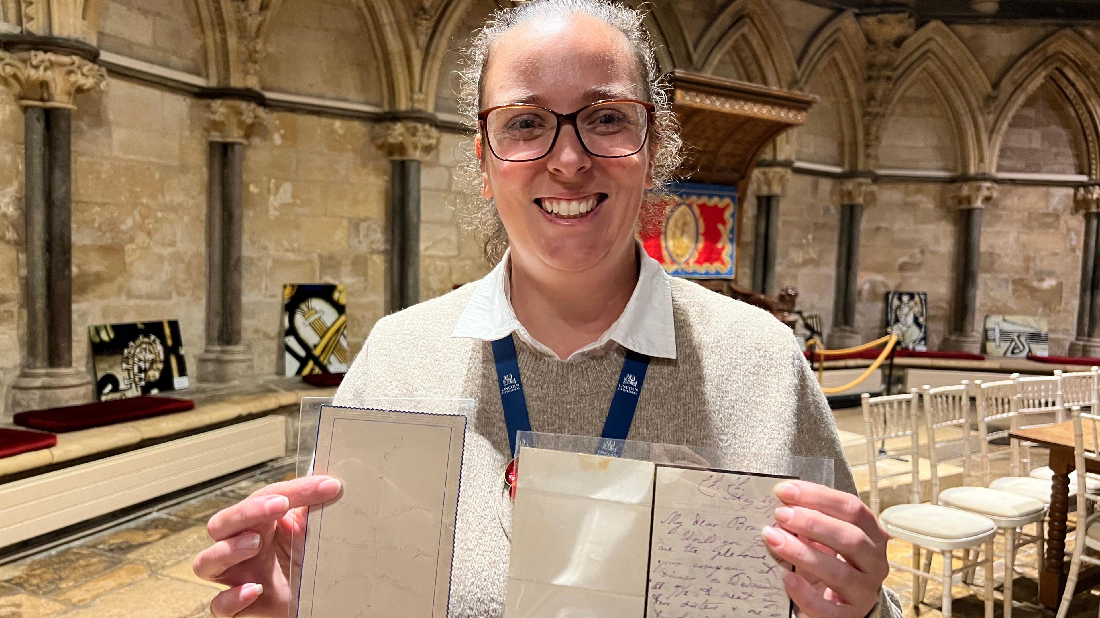 A woman standing inside Lincoln Cathedral holding two framed documents, one with handwritten text and the other with faint markings. There are arched columns in the background.