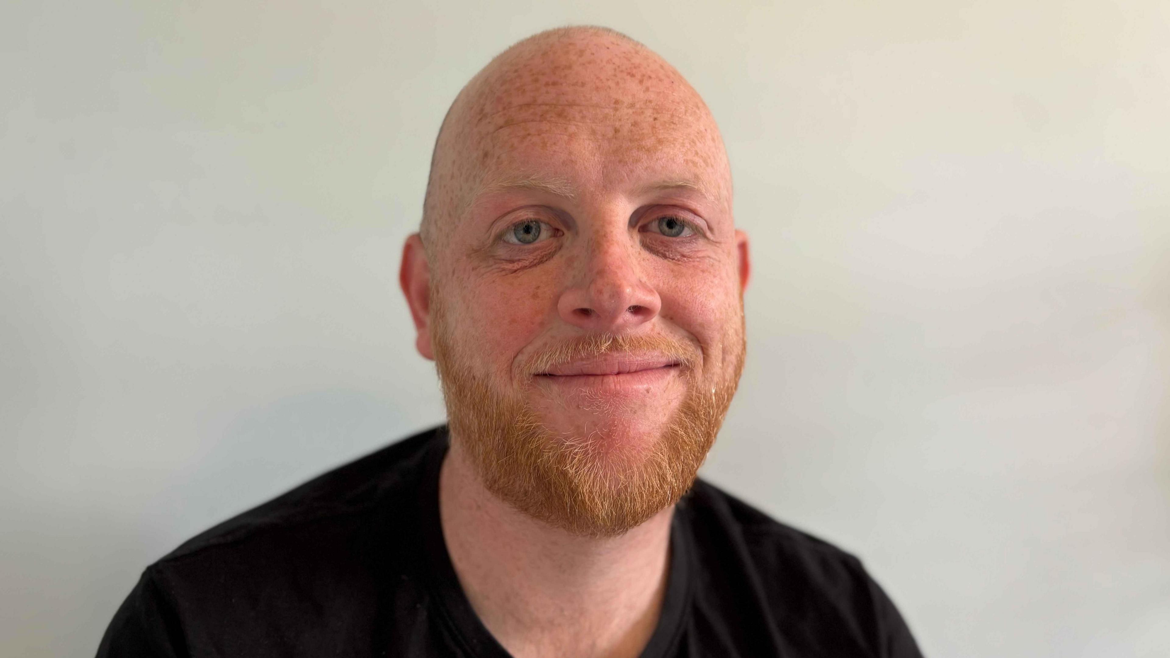 Liam Smith is looking towards the camera with a light smile. He has a short ginger beard, and is wearing a black T-shirt.