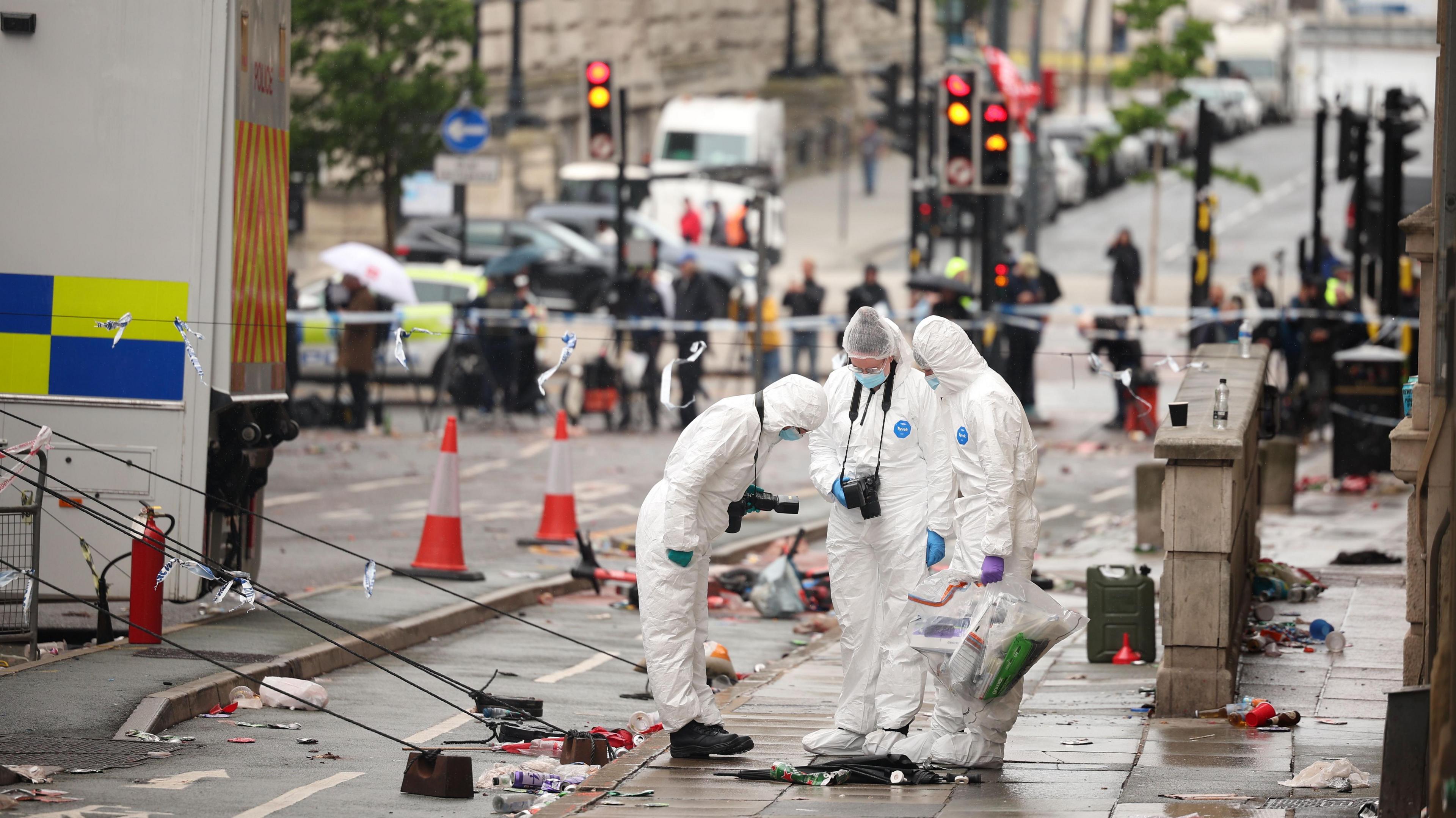 Three people are wearing forensic suits with cameras around their necks. They are standing on a pavement with rubbish scattered around them. In the background cars are parked on the road.