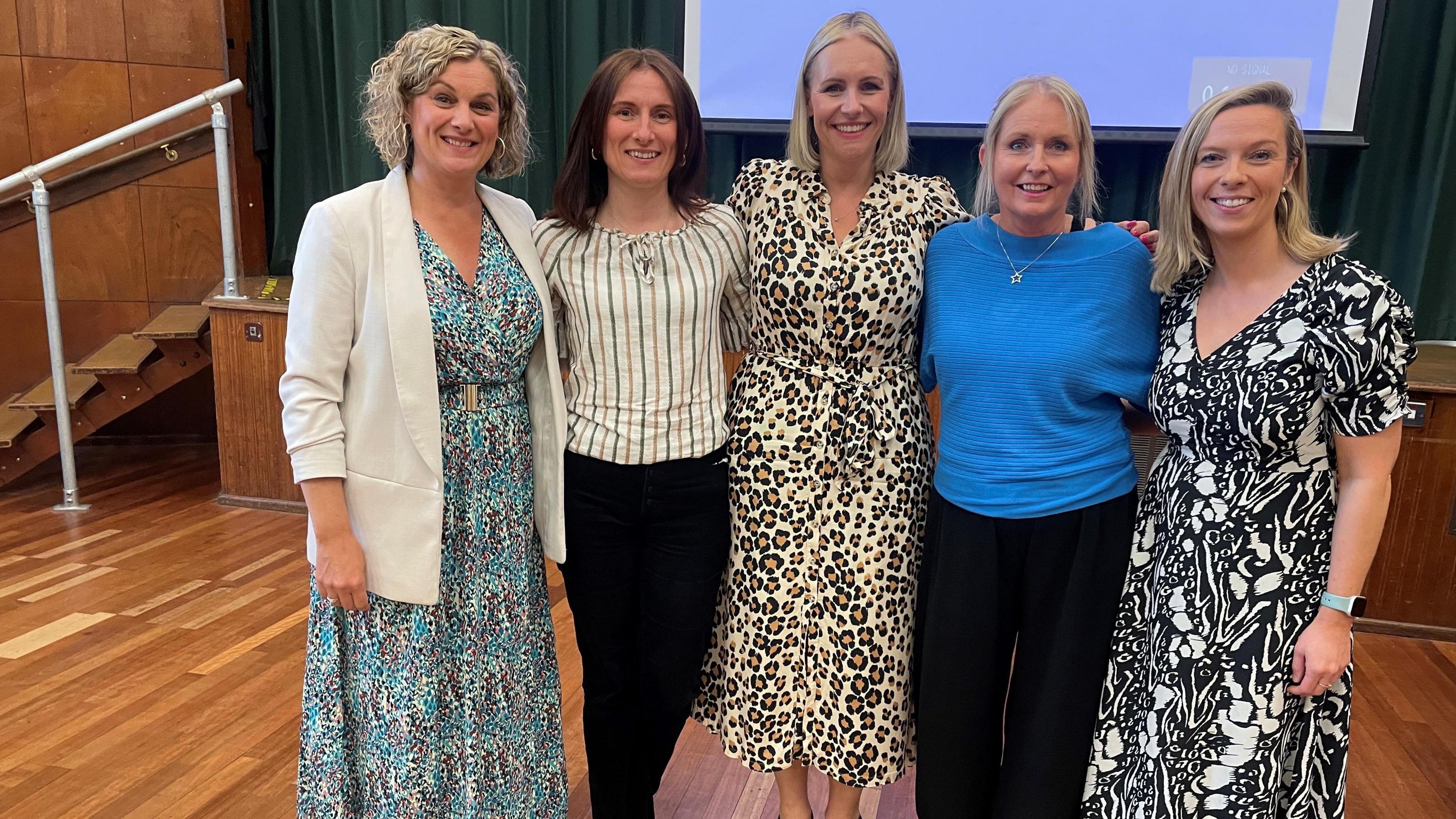 A group of five women stand in an assembly hall with their arms around each other. Three are wearing patterned dresses and two are wearing dark trousers and a top. All women are smiling into the camera.