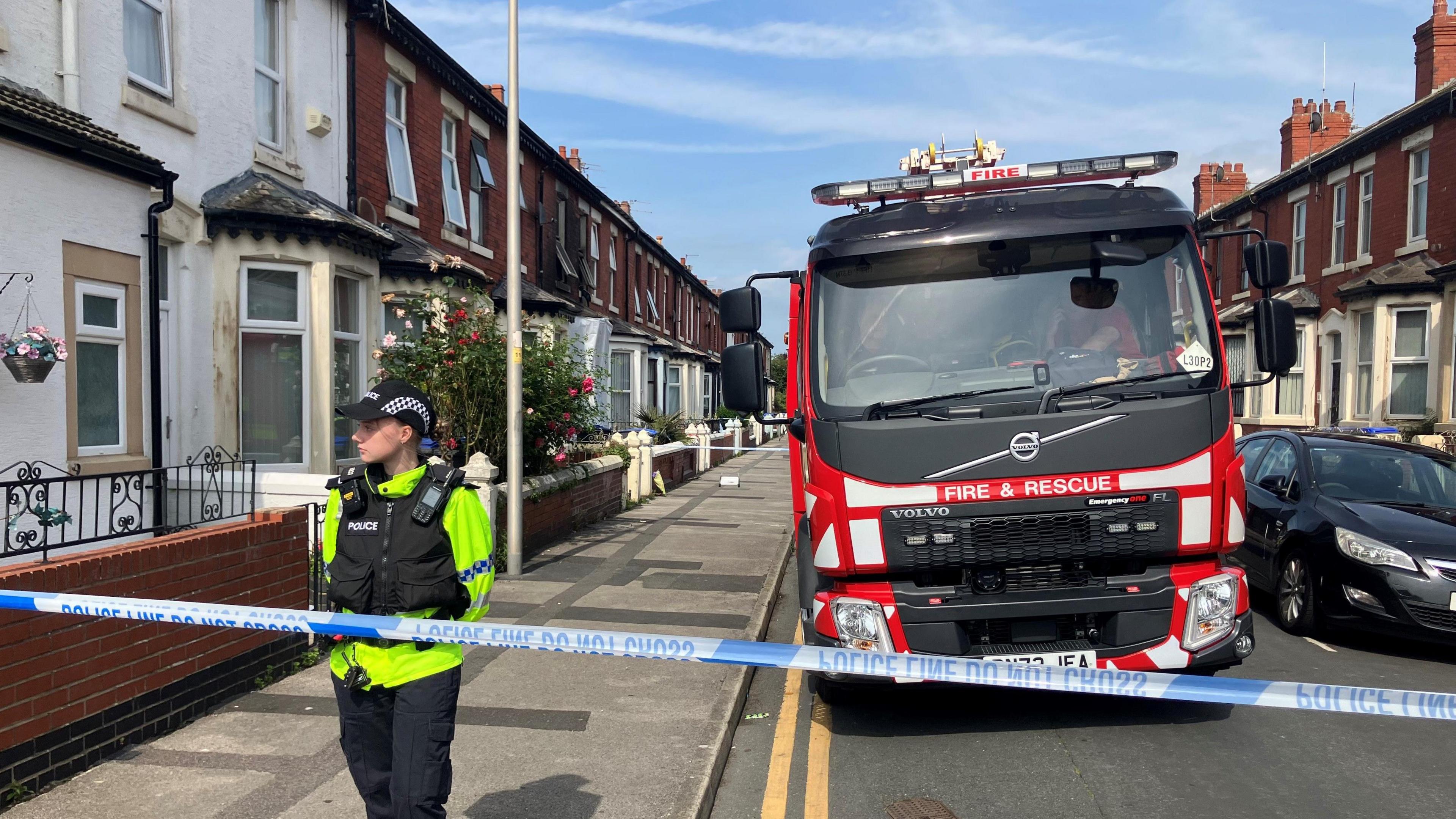 A police officer stands outside the scene of a house fire as a fire truck is seen behind him on the street. Police tape crosses the front of the pic. The houses are Victorian in style with big bay windows. 