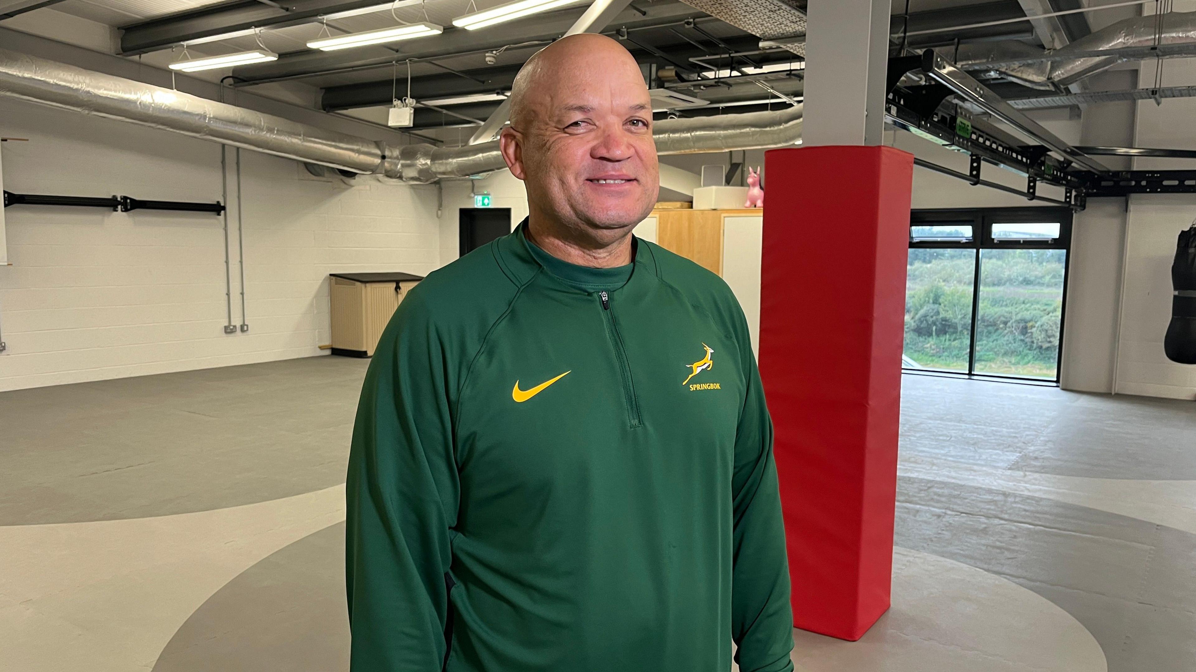 Deon Davis wearing a green Springbok jumper. The jumper has a yellow Nike tick on the left and a Springbok logo on the right. He is smiling towards the camera. In the background is a red and white pillar and white walls. 