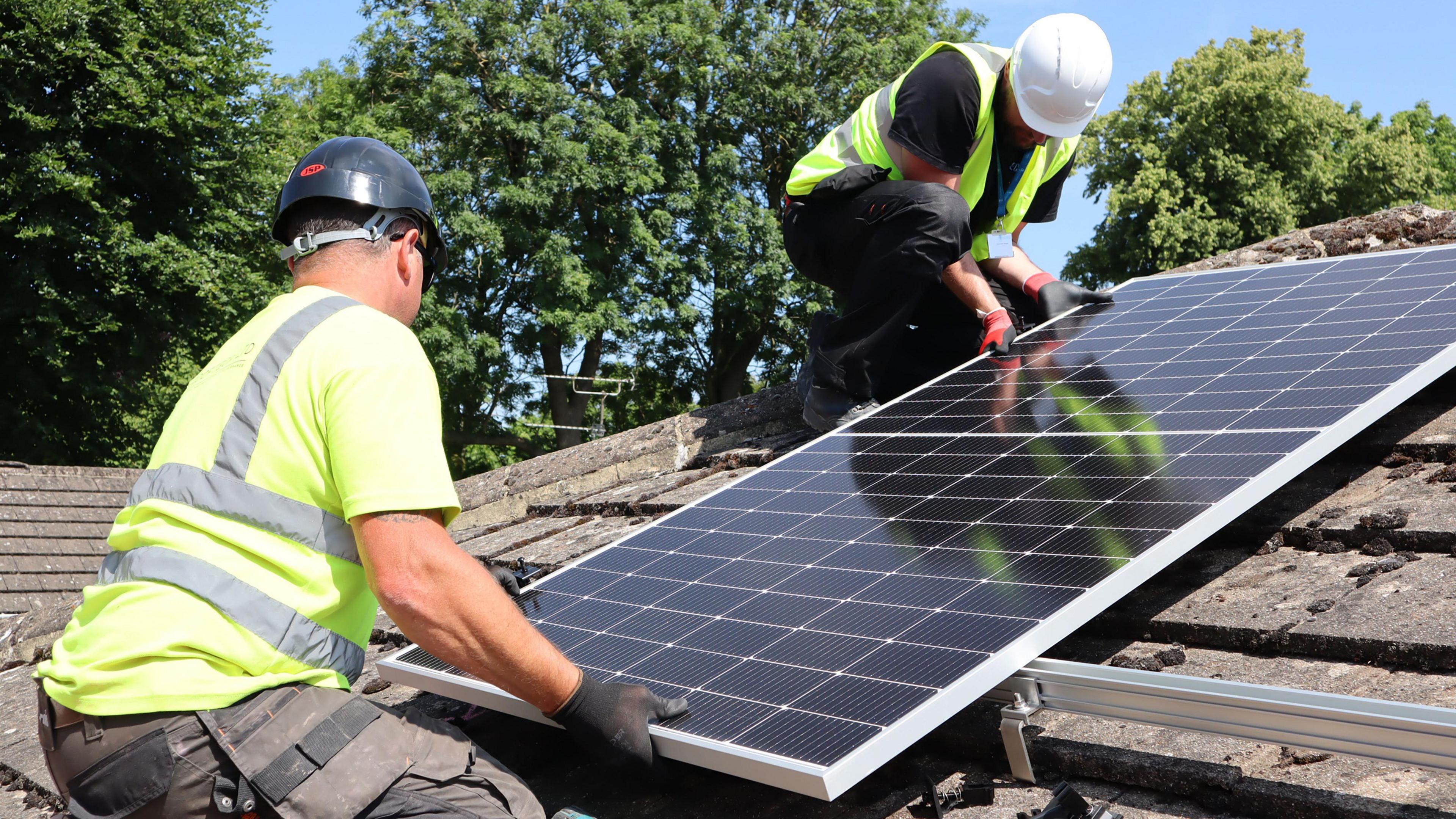 Two men in high vis vests and hard hats lifting a solar panel onto a roof