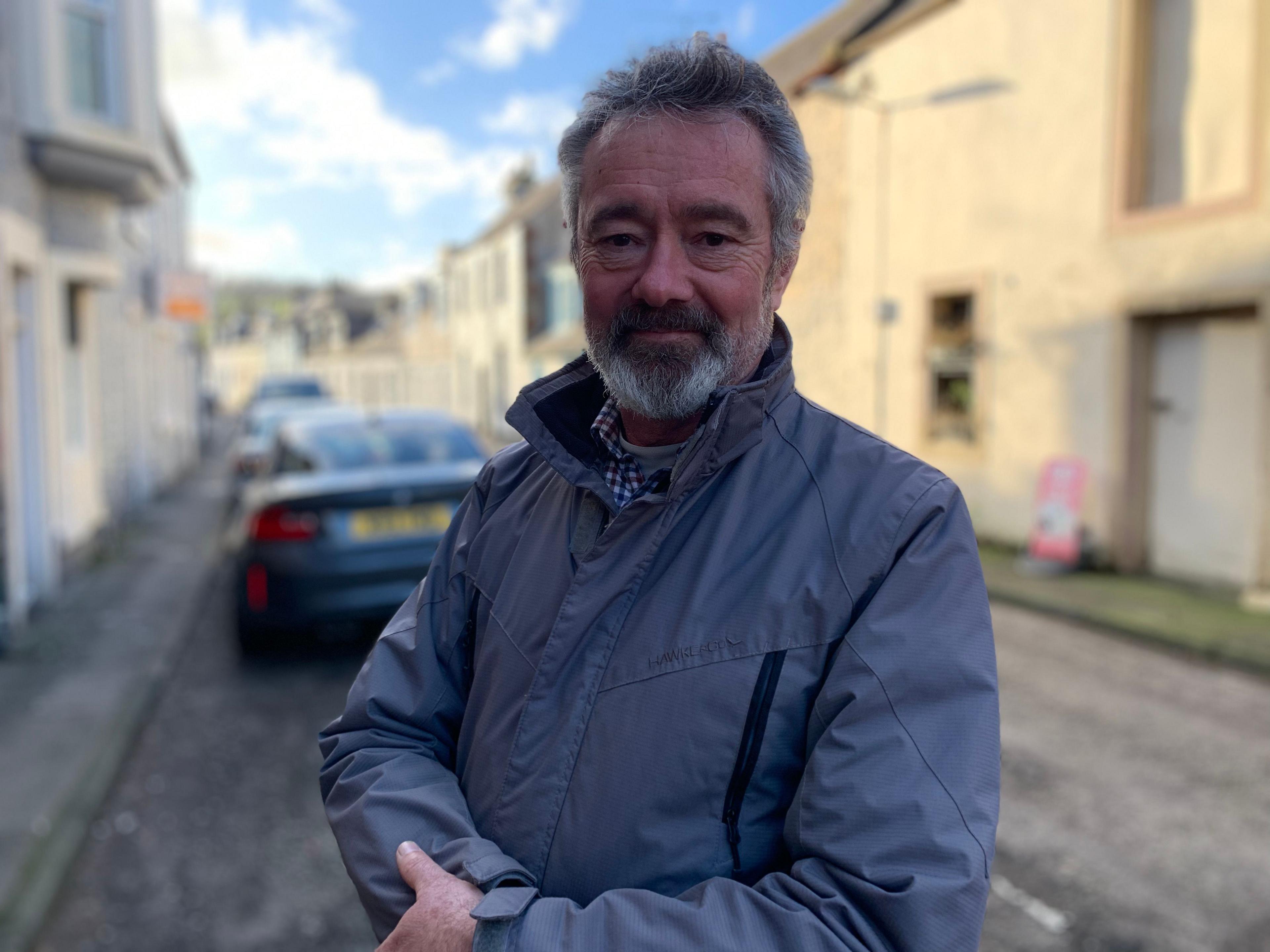 A grey haired man with a beard in a grey jacket with a checked shirt underneath stands in a side street in Moffat with cars parked behind him