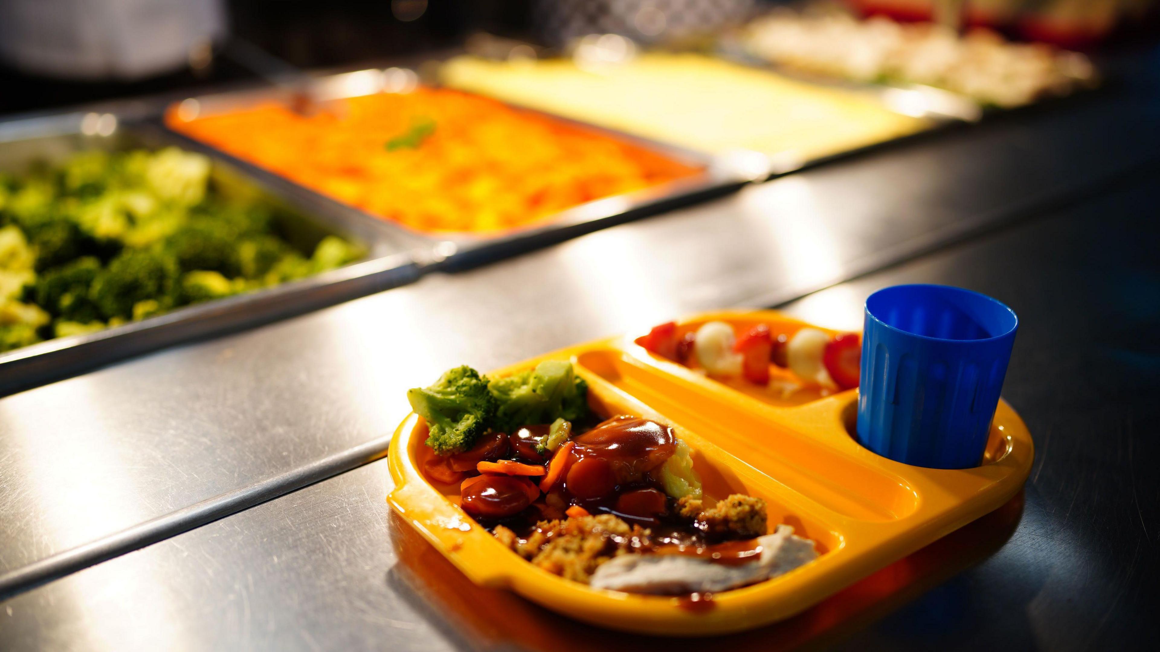 A yellow school lunch tray filled with chicken and vegetables, fruit and a blue plastic cup. Larger trays of broccoli, carrots and potatoes are out of focus in the background.