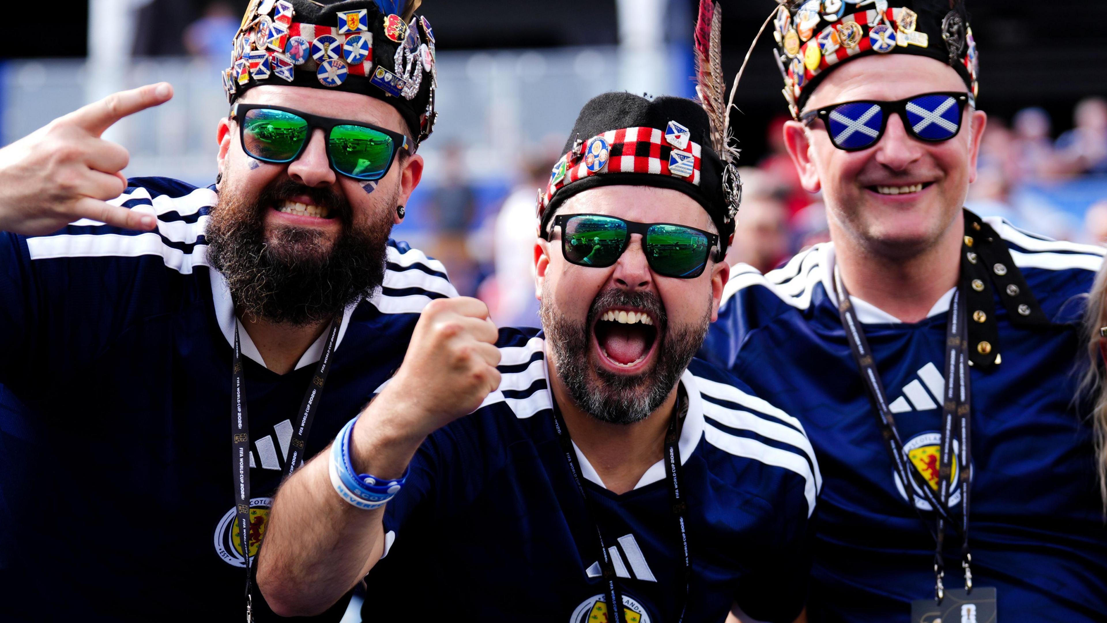 Three Scotland fans wearing blue home tops cheer at the camera. They are wearing Scottish hats and sunglasses