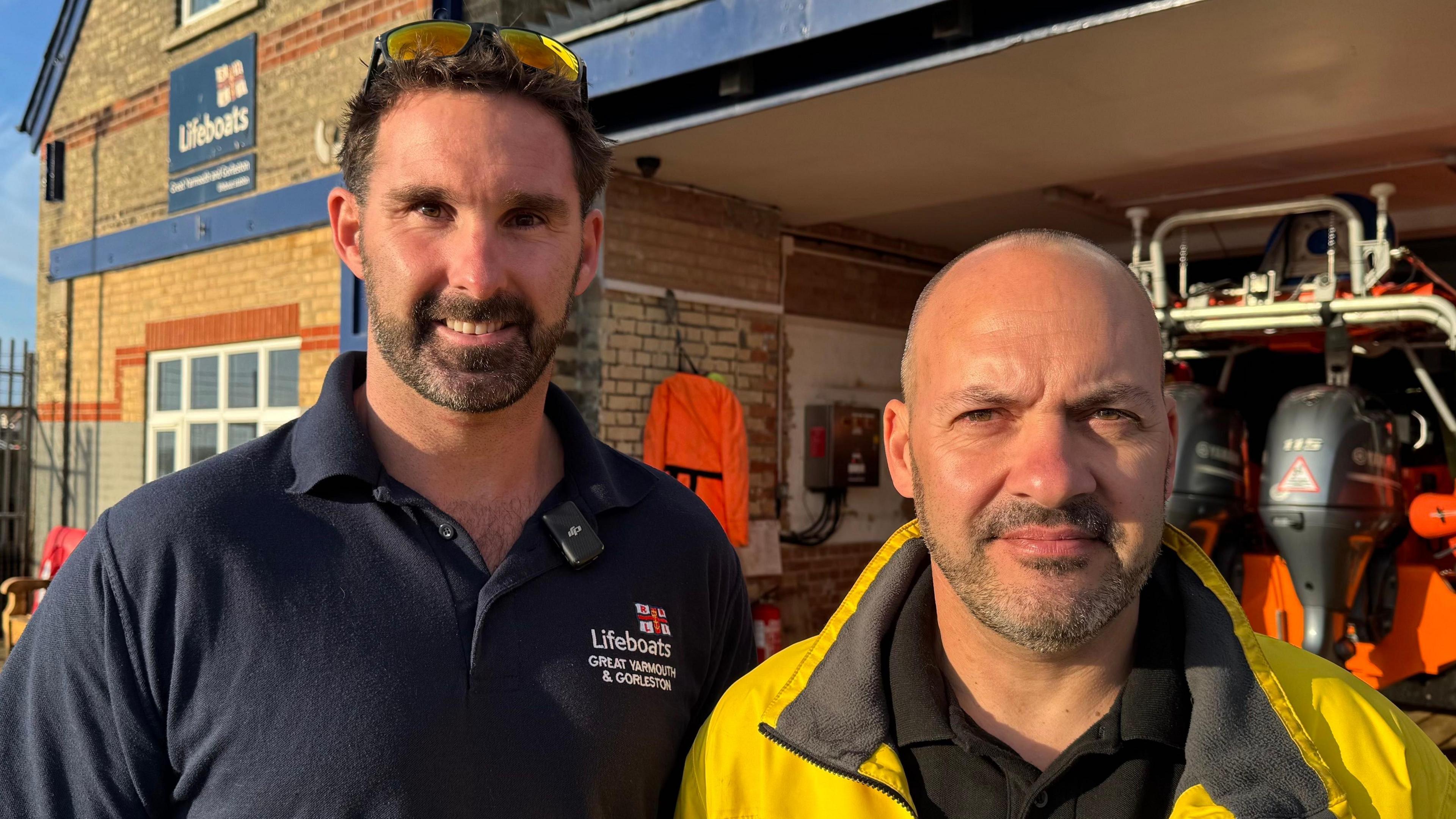 John McLennan is wearing a navy blue polo shirt and has short brown hair and is looking towards the camera smiling. Ben Penning is standing on the right and is wearing a yellow jacket, a black polo shirt. Behind them is a RNLI lifeboat. 