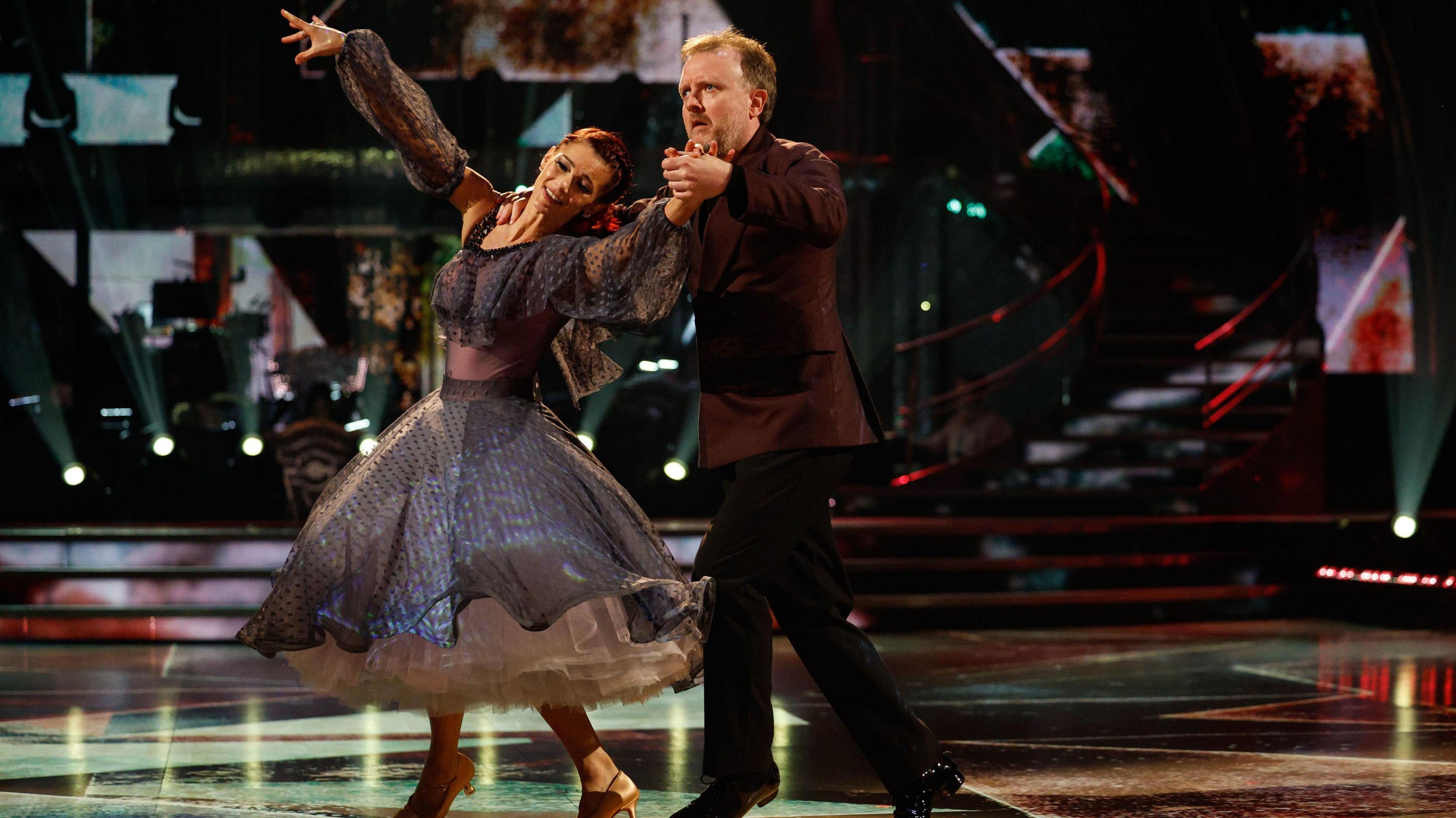 Chris McCausland and his Strictly Come Dancing partner Dianna Buswell dance on the set of the programme. Dianne stands in front of Chris with her back to him and her left hand raised whilst holding his right hand. She wears a dark purple dress and heels. Chris has his right hand on her shoulder and wears a brown shirt with black trousers and shoes.