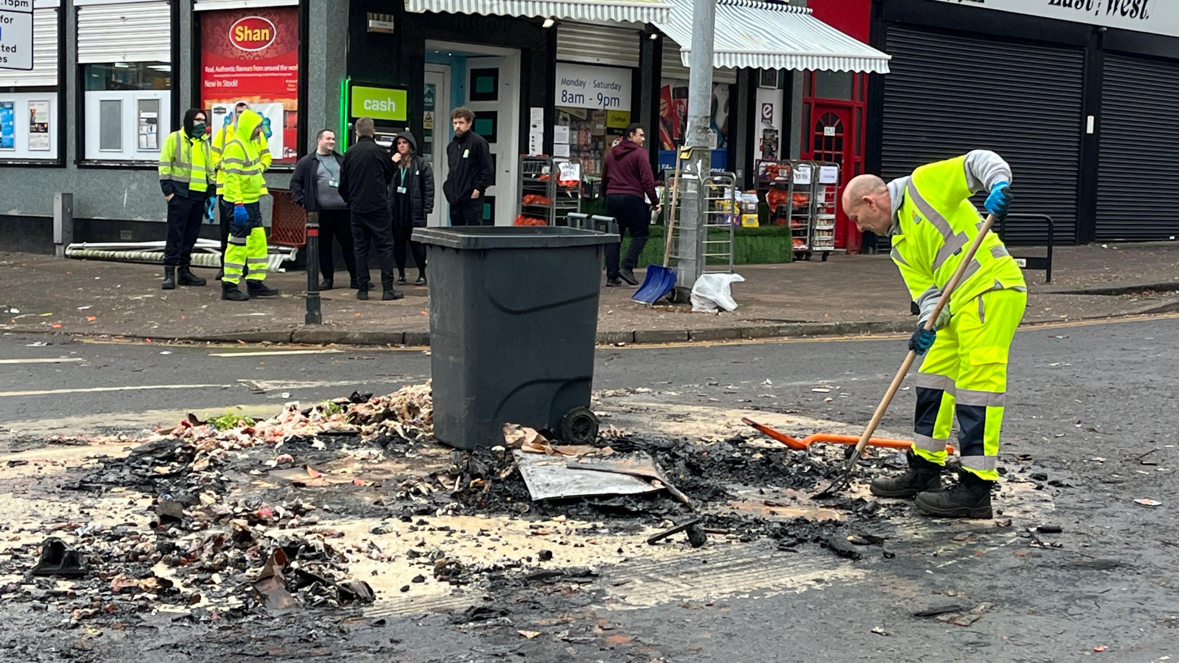 A shot of Albert Drive in Pollokshields - a council worker clears blackened debris from firework disorder into a large grey bin. A row of shops is visible in the background with a number of people standing outside.