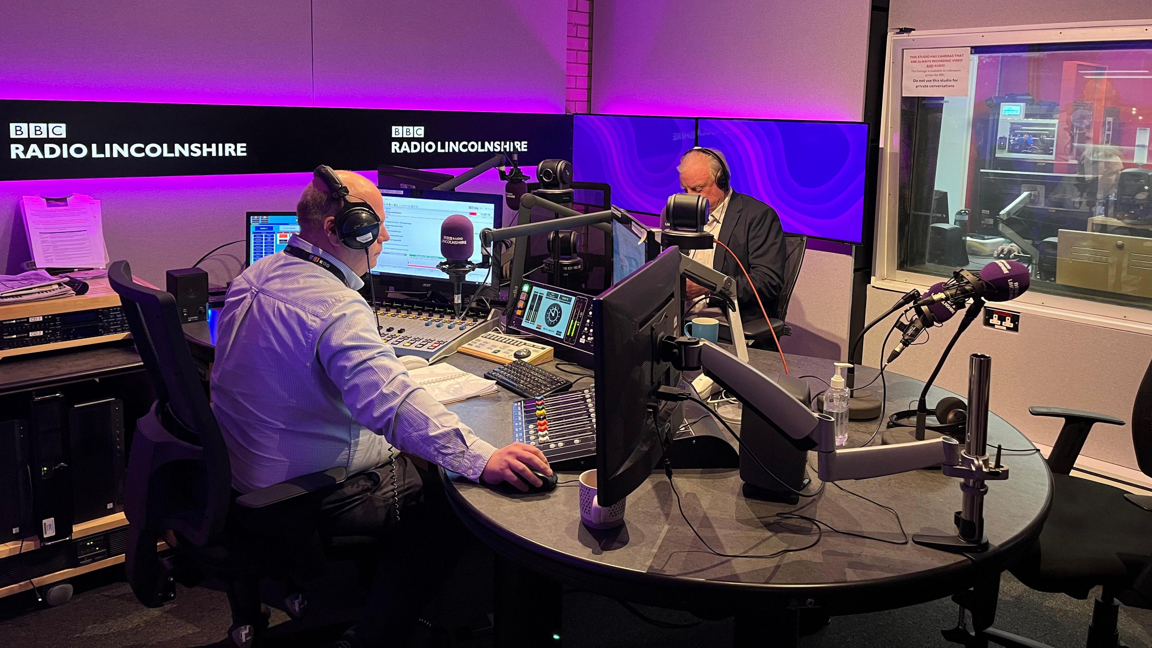 Sean Dunderdale, a man wearing a light-coloured shirt sits behind a radio system. Facing him is Sir Edward Leigh, wearing a white shirt and a navy blazer. They are in a radio newsroom, surrounded by radio equipment.