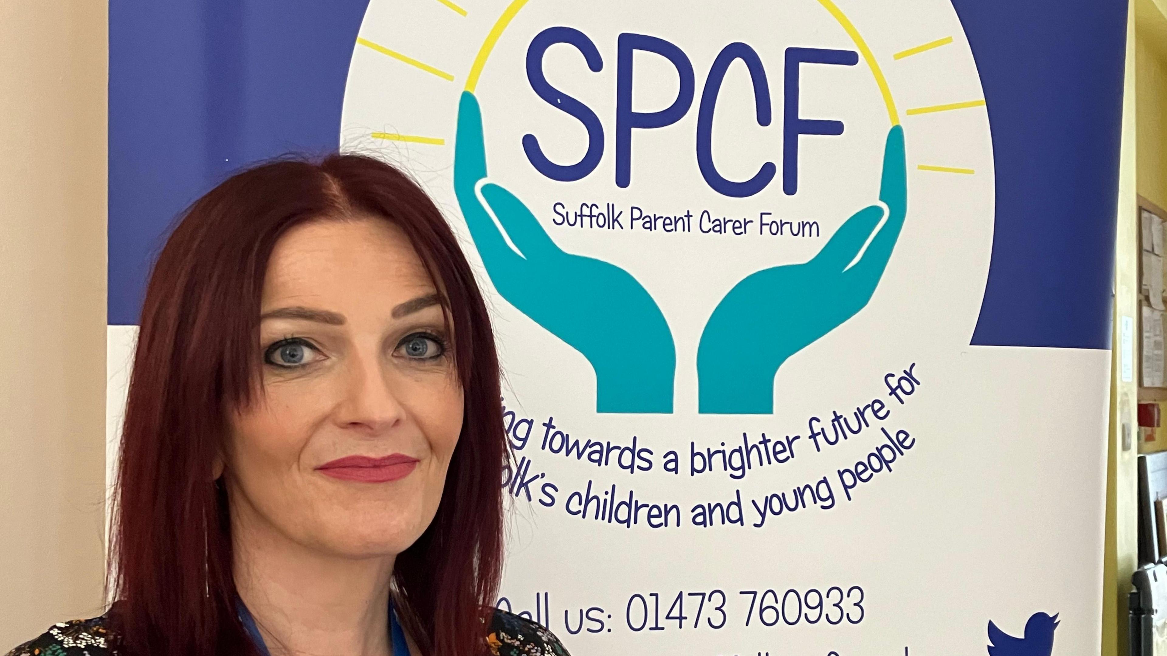 A woman with dark red hair standing in front of a signing displaying the Suffolk Parent Carer Forum logo