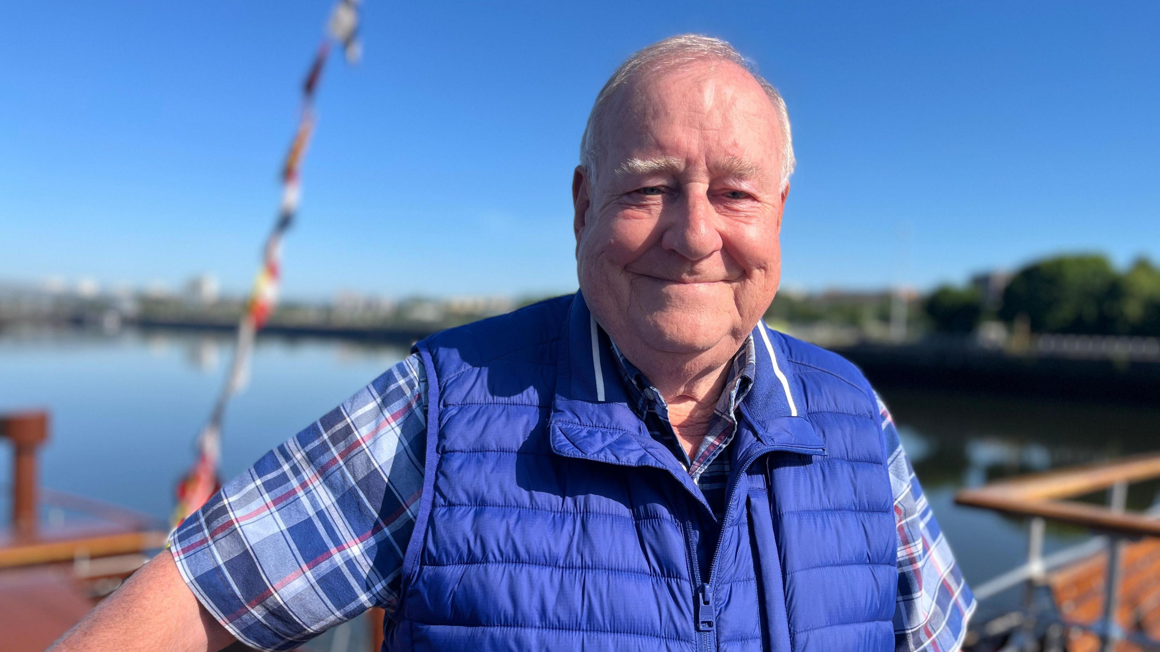 Douglas McGowan, a smiley man with white hair, poses on the Waverley, blue skies and water behind him. He is wearing a blue checked shirt and a royal blue gilet.