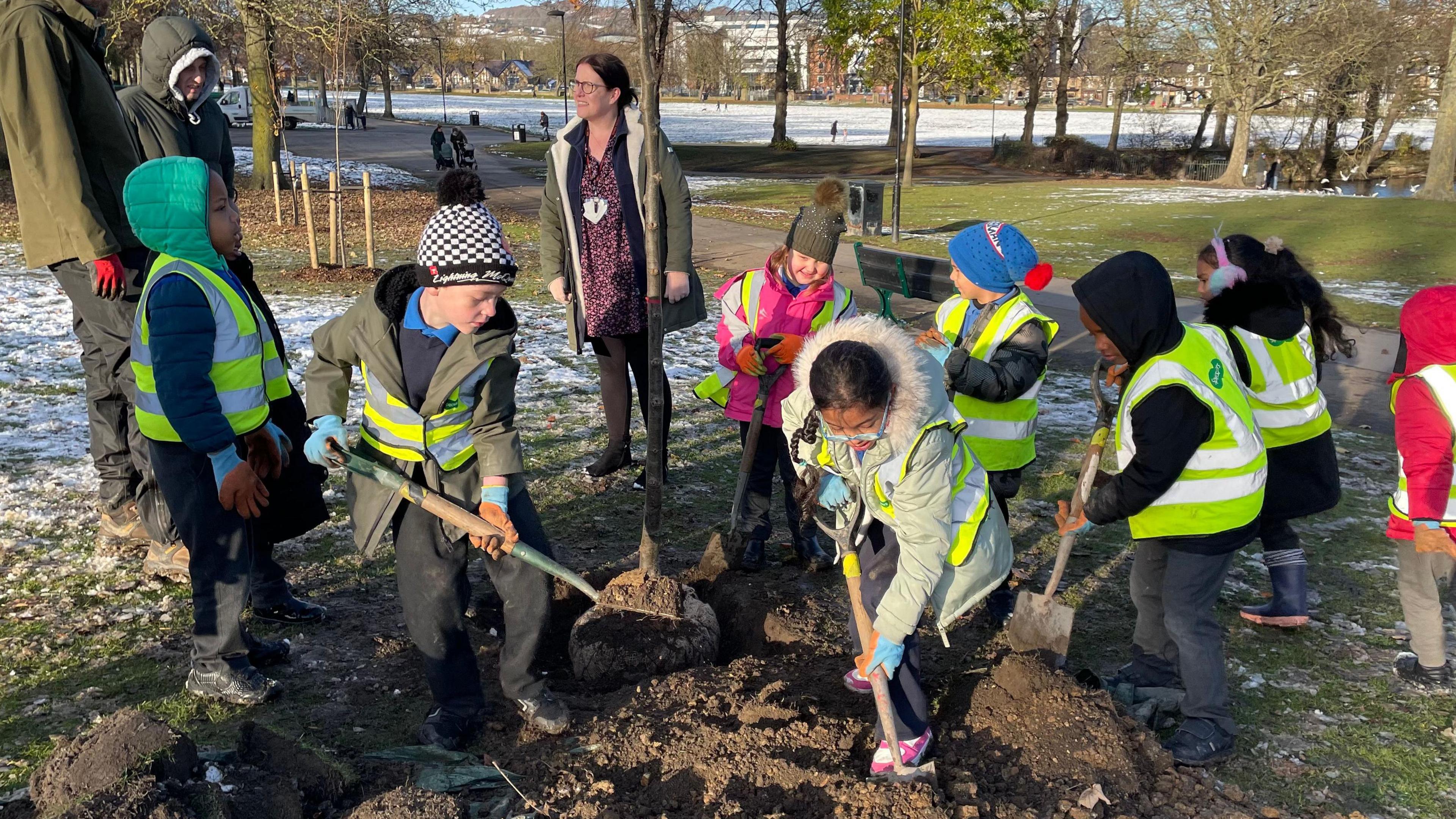 Young children are wearing high-visibility jackets and using small shovels to fill in a hole that's been dug in the ground. In the hole is a tree sapling. There's snow on the ground around them.