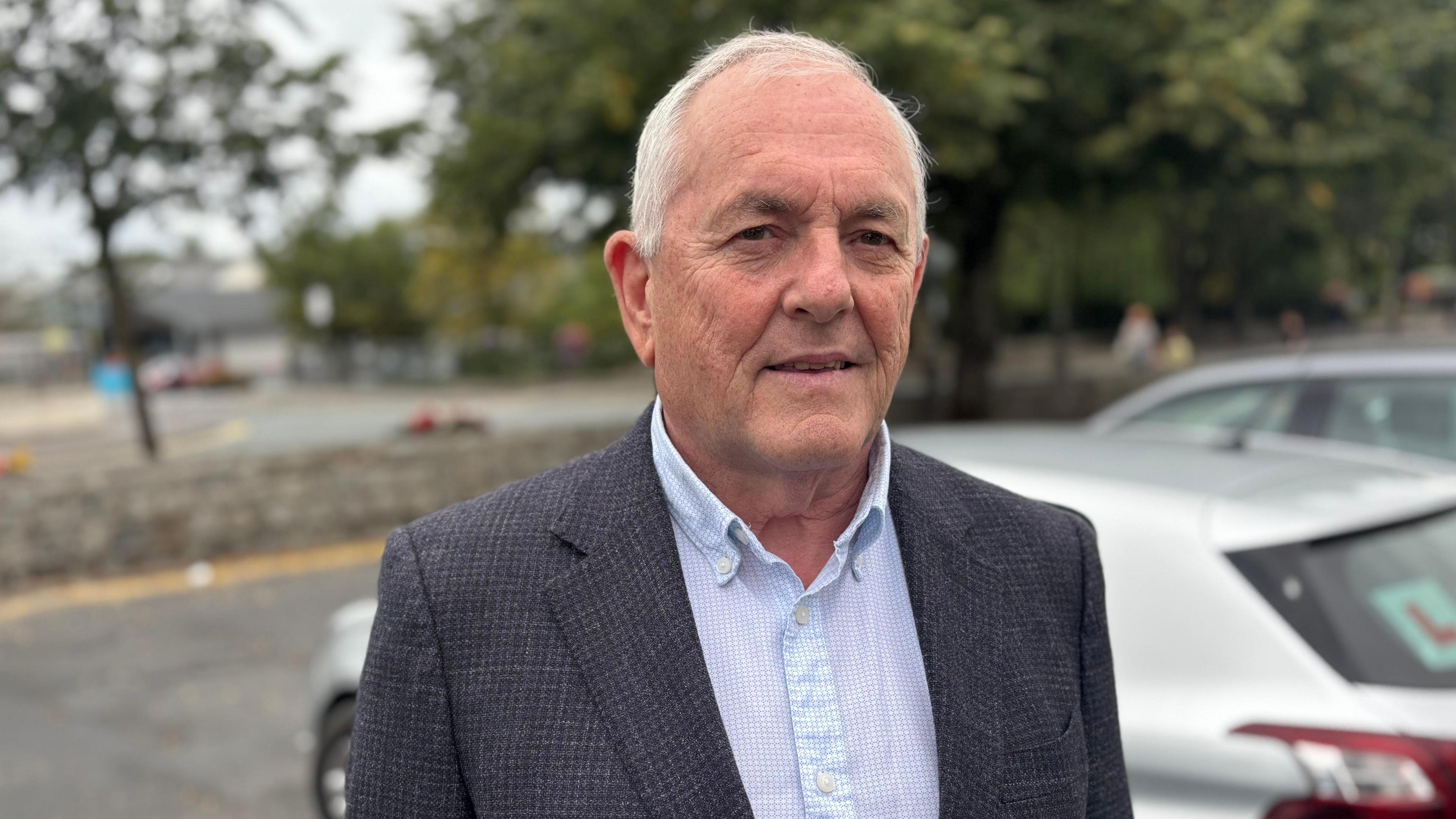 Fianna Fáil TD John McGuinness, with short white hair, wearing a blue shirt and dark grey suit. There is a silver car parked behind him.
