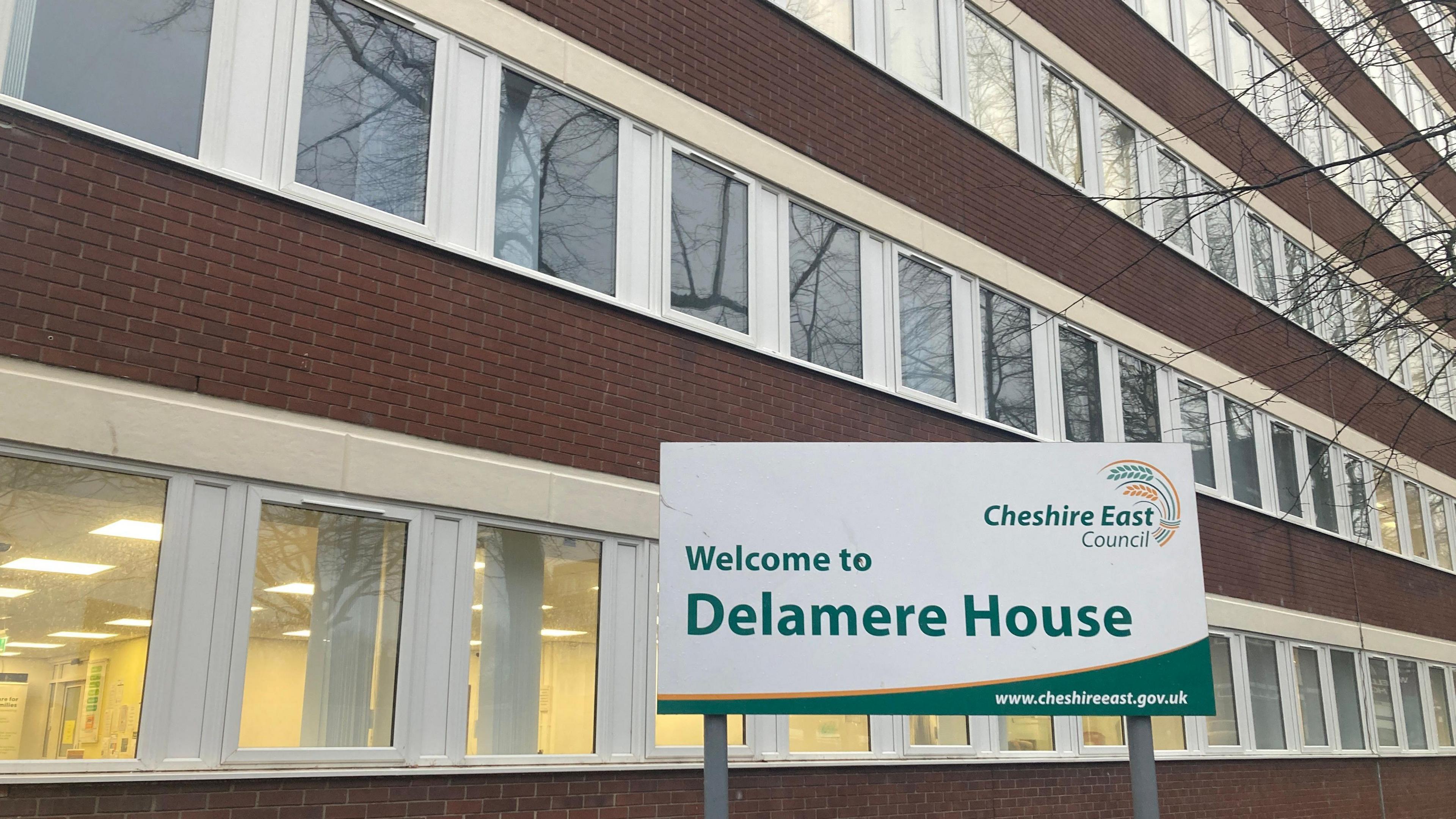 Cheshire East Council's headquarters at Delamere House in Crewe. It is a brown brick buildings with white plastic window surrounds all the way along the ground floor and first floor. There is a sign outside that says Welcome to Delamere House.