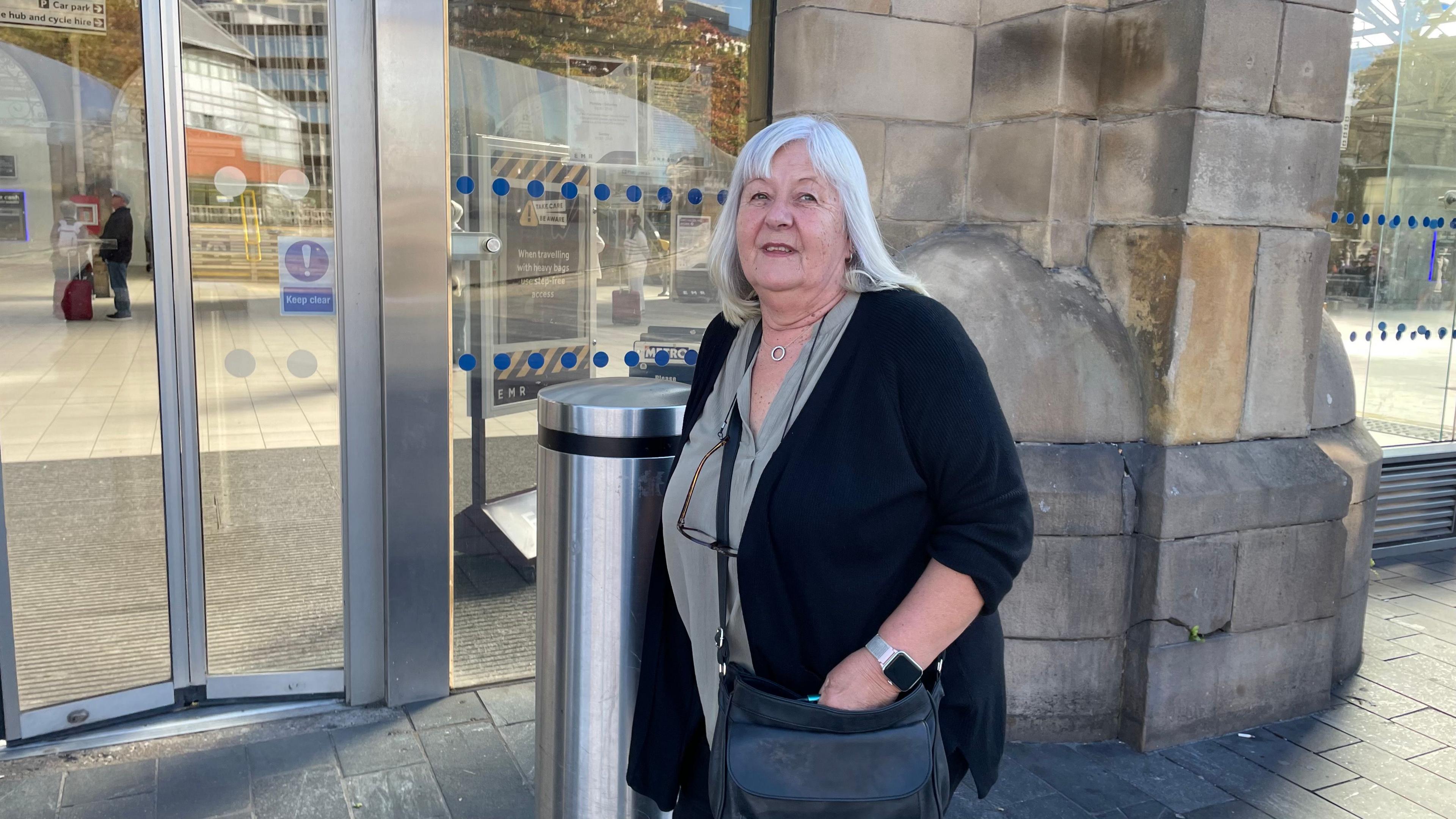 A woman in her sixties stands outside a railway station. She is wearing a black cardigan and black handbag.