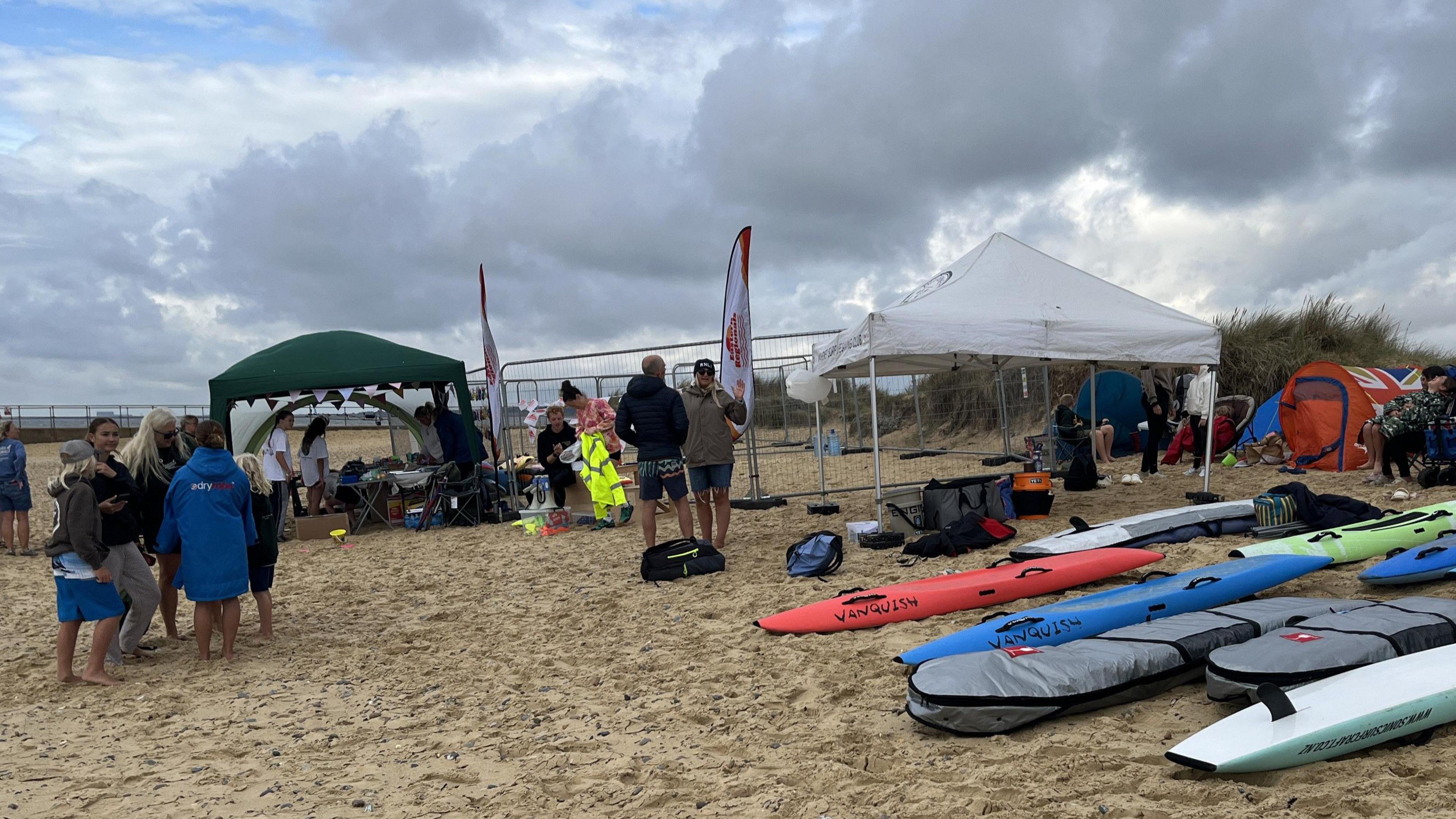 Body boards rest on a beach while people mingle together nearby and under gazebos that have been set up.