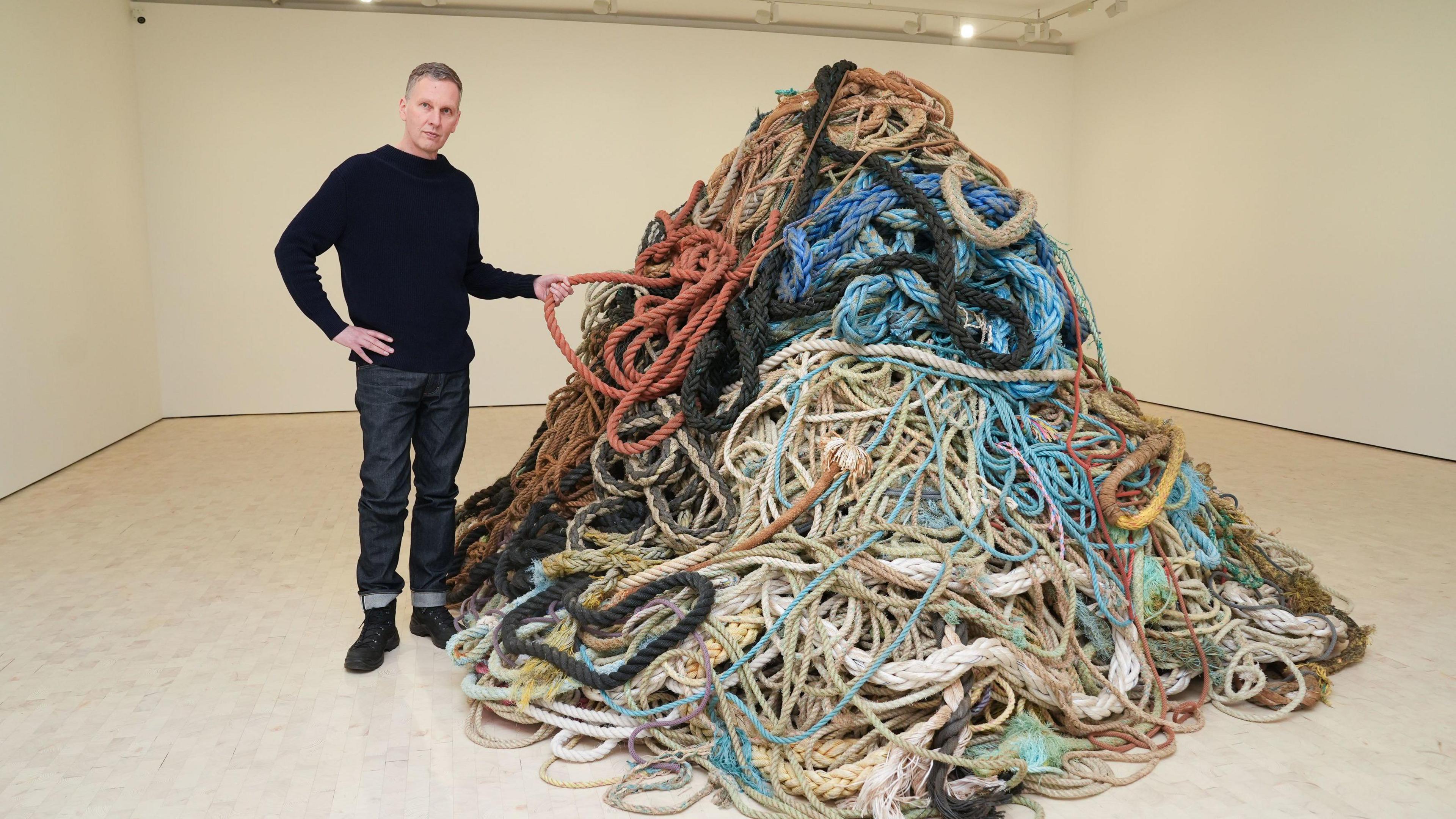 Artist David Shrigley stands next to a pile of rope in an art gallery