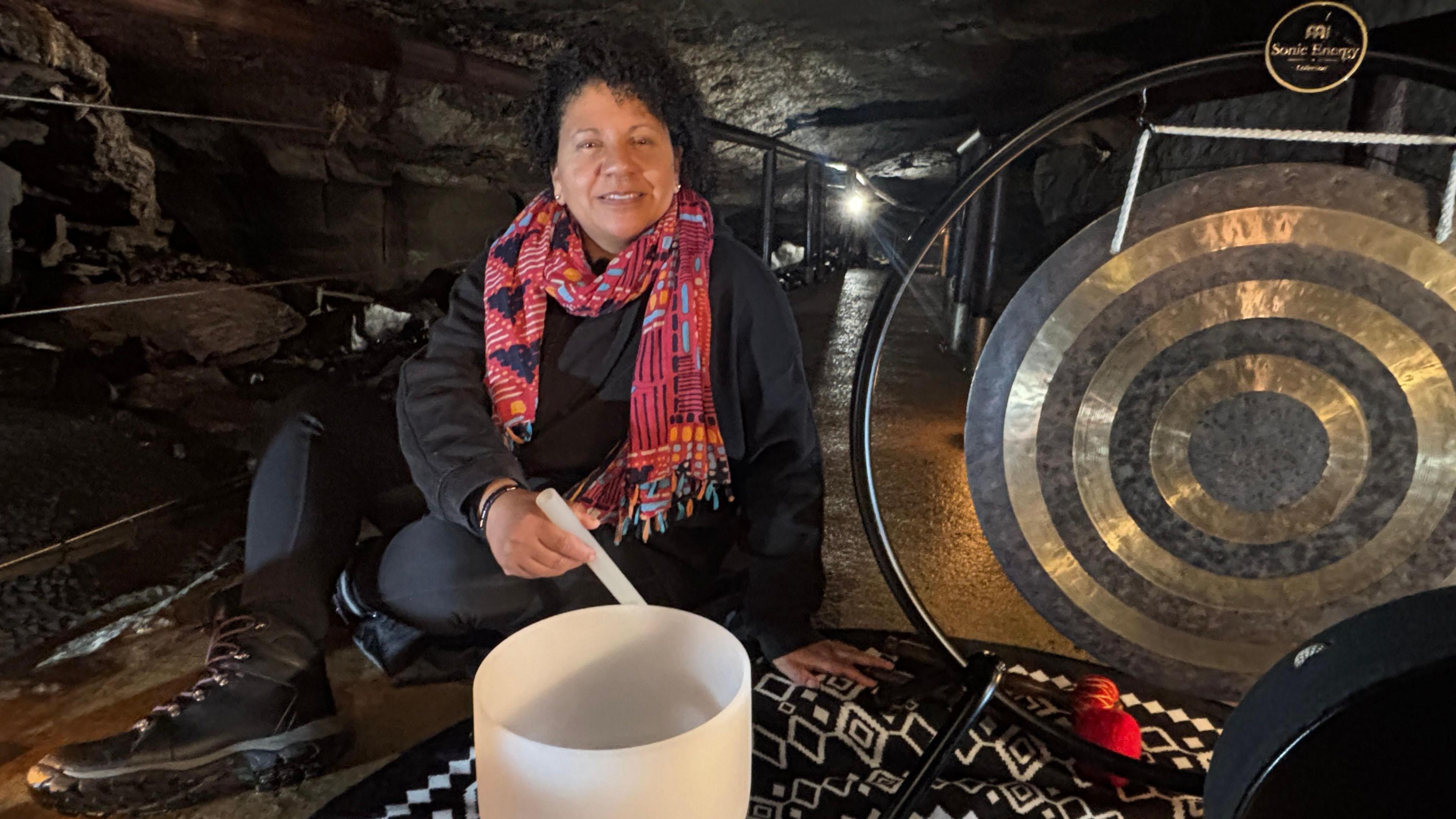 A woman sits in an underground cave. She has a large crystal bowl in front of her and a metal gong beside her. She is dressed in black and wears hiking boots. She has a colourful scarf and is smiling as she touches the rim of the crystal bowl with a crystal stick.