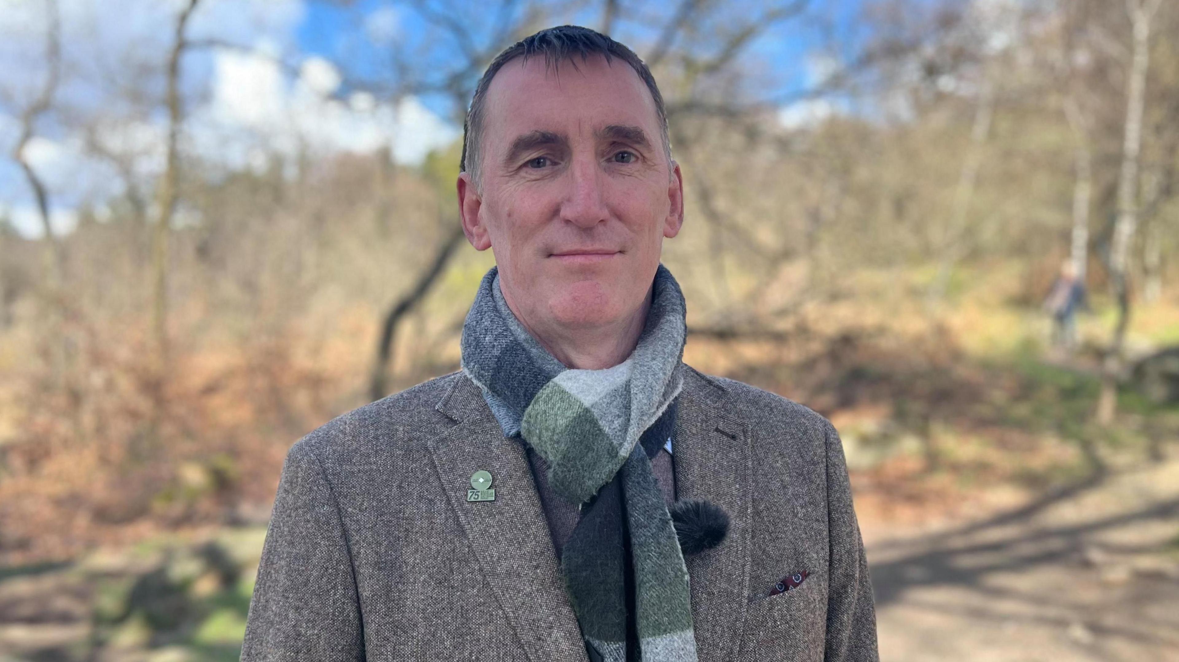 Man in grey tweed jacket stood infront of forest landscape