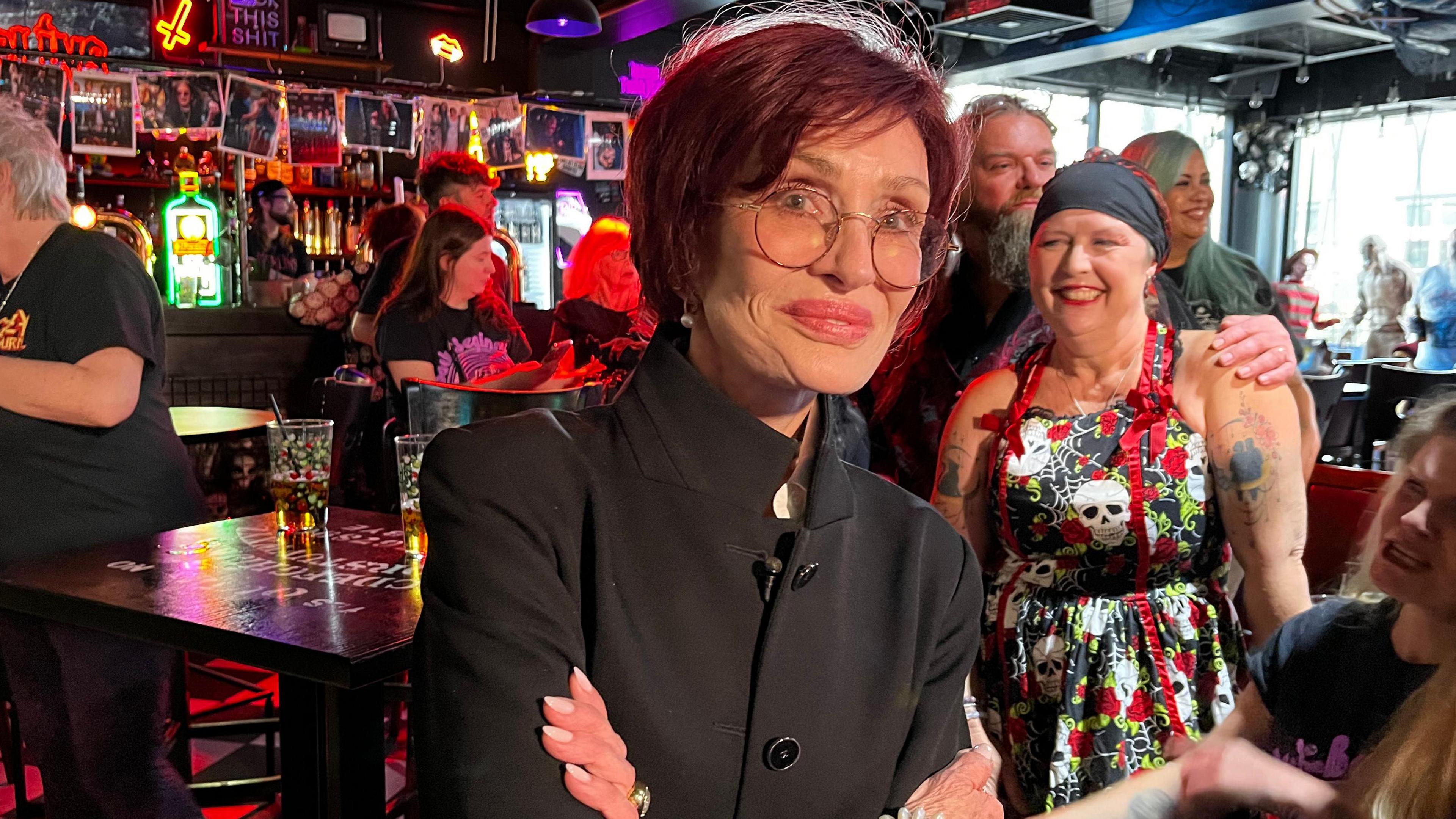 Sharon Osbourne wearing a black suit with a group of smiling people in the background inside a bar.