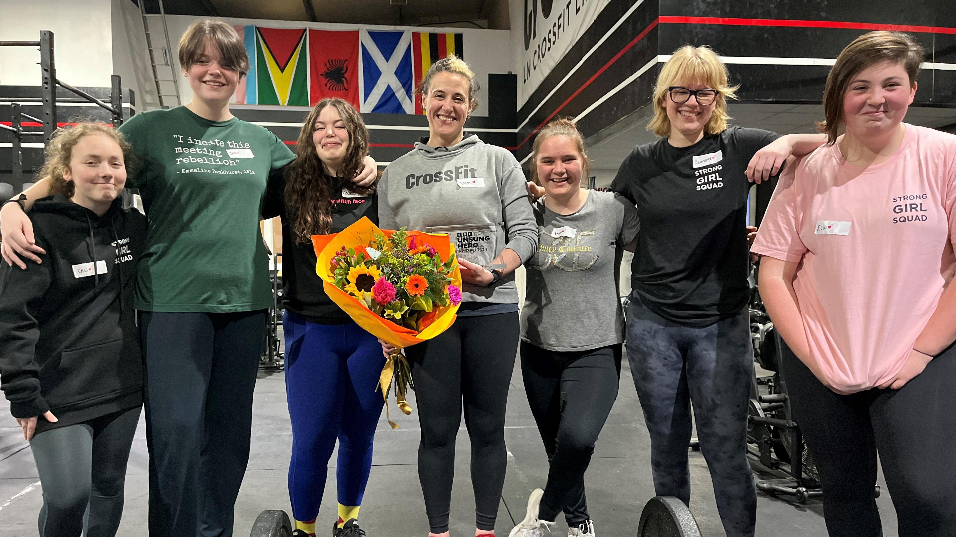 Kristen Ingraham-Morgan stands in the centre of a row of seven women and girls, all wearing gym clothes and some with shirts marked "Strong Girl Squad". Ms Ingraham-Morgan is holding her BBC Sports Personality Unsung Hero trophy  and large bunch of colourful flowers. They are in a gym, surrounded by weights, a rowing machine, and an exercise bike.