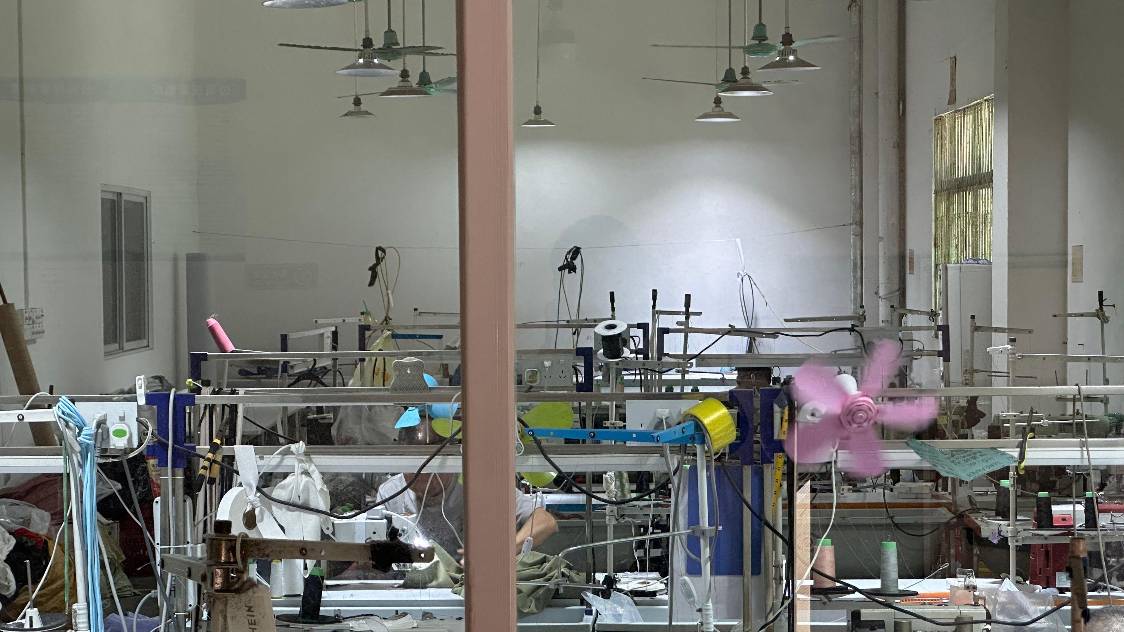 A window with a brown sill look into a room with sewing machines with a row of lights and green fans. Another purple fan can also be seen lower down. 