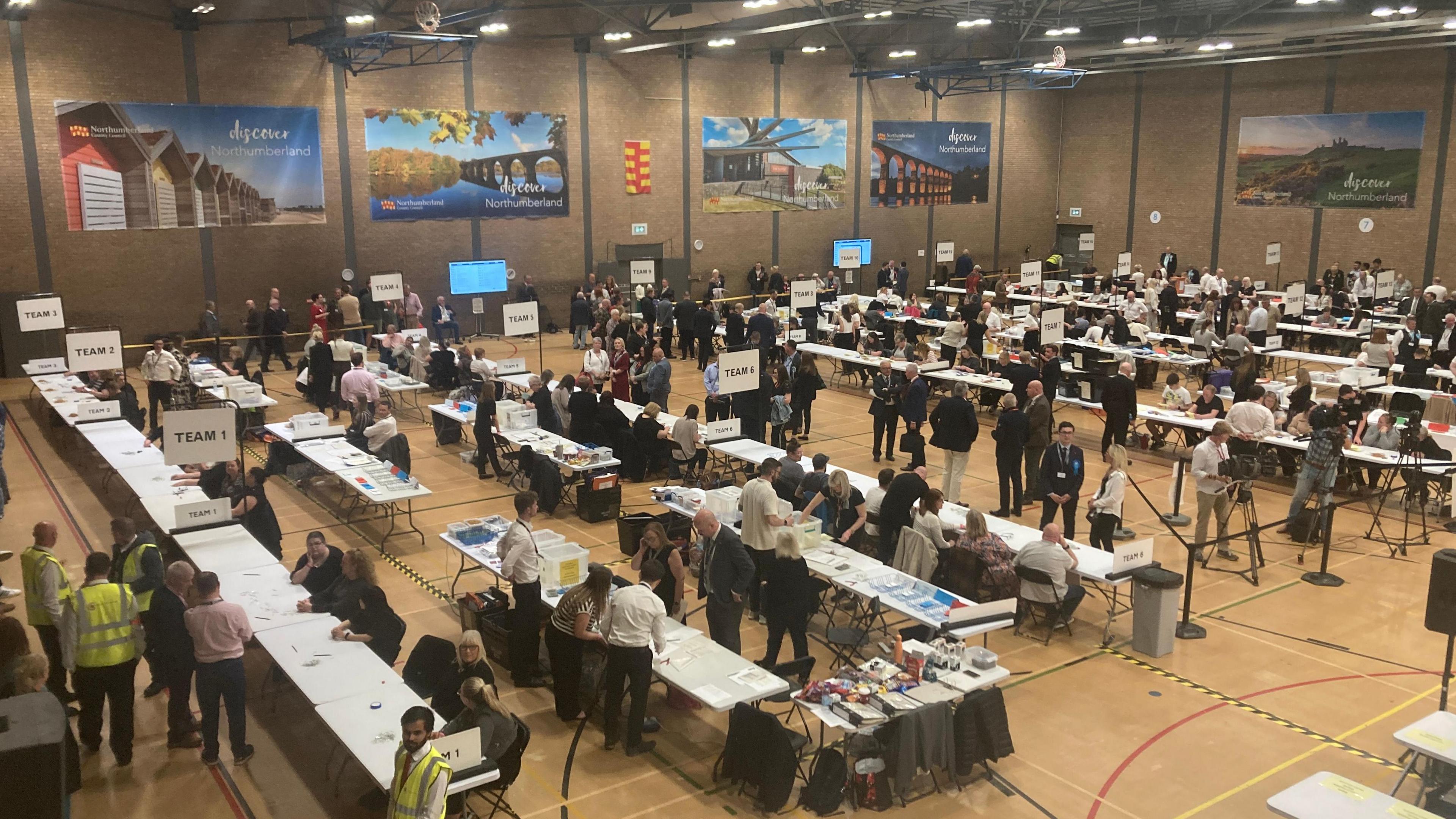 Votes are counted in a large hall. Several rows of tables are laid out with volunteers counting ballots. A number of banners advertising Northumberland decorate the walls.
