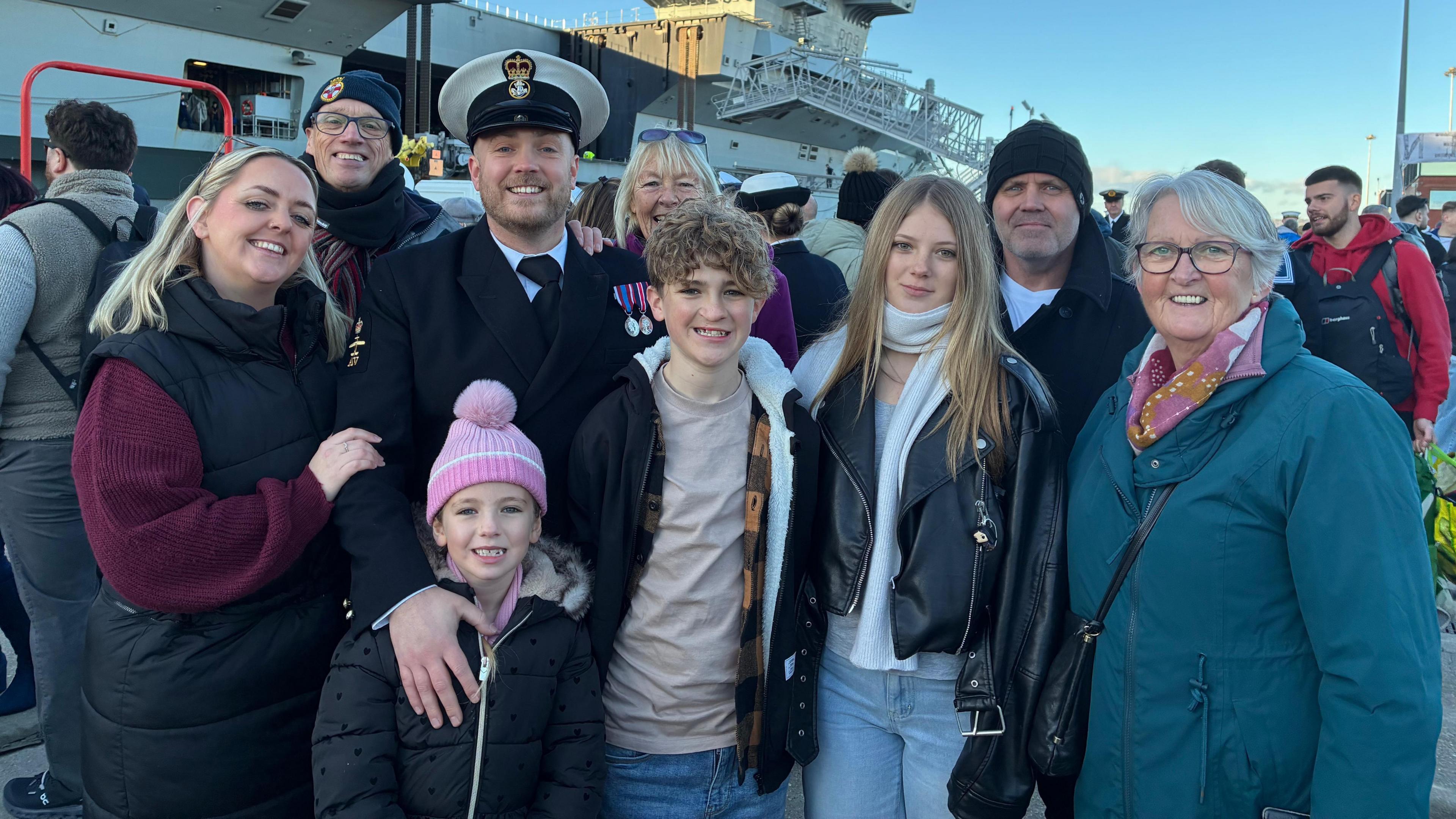Andrew 'Bouncy' Ball wears a Royal Navy suit and hat, with medals on the left panel of his jacket. He is standing with his three children and other loved ones all smiling at the camera.