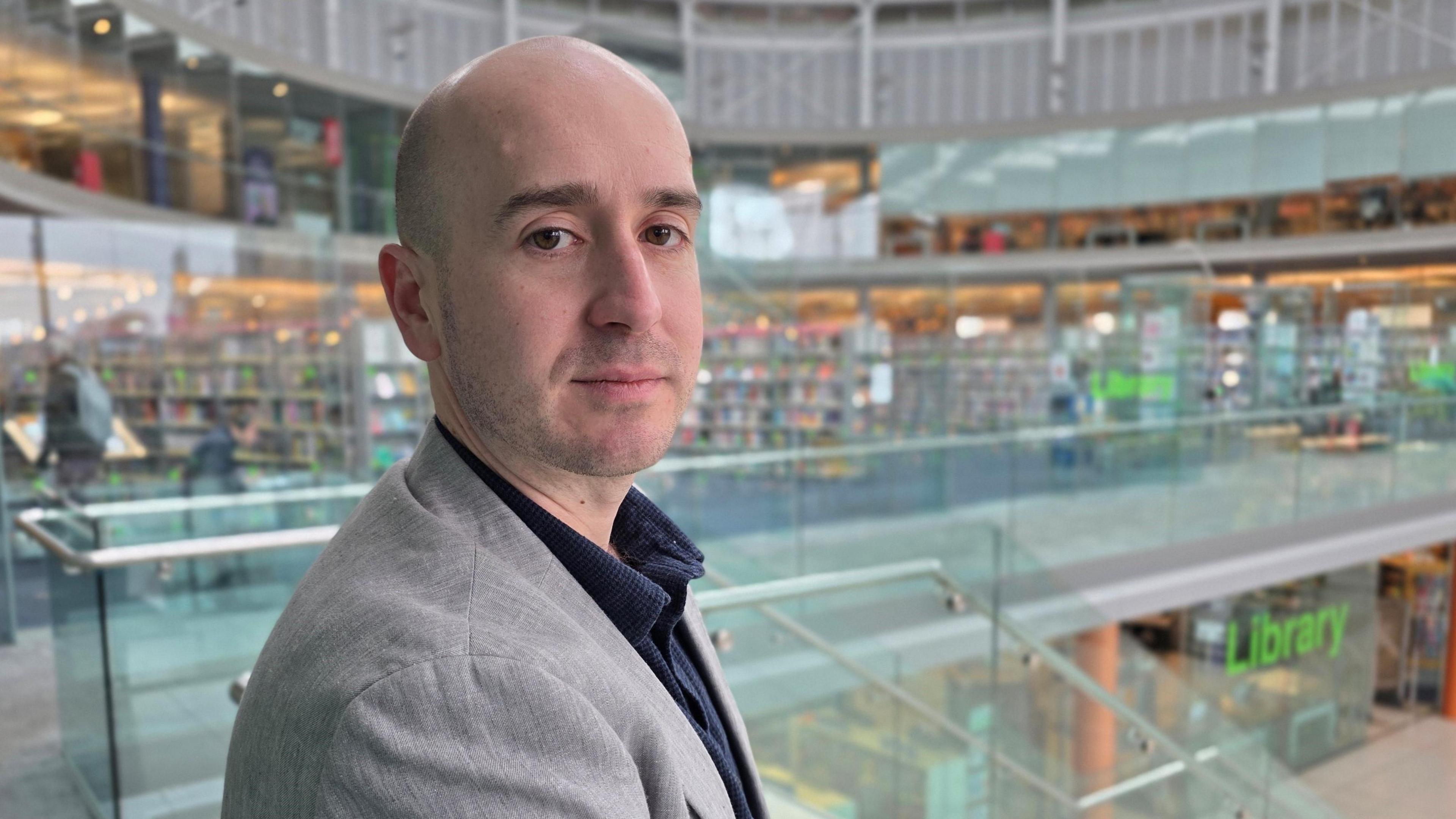 Ben Goldsborough is looking at the camera over his right shoulder. He has a grey suit jacket on over a blue shirt. He is standing on a glass internal balcony, with the library visible behind him.