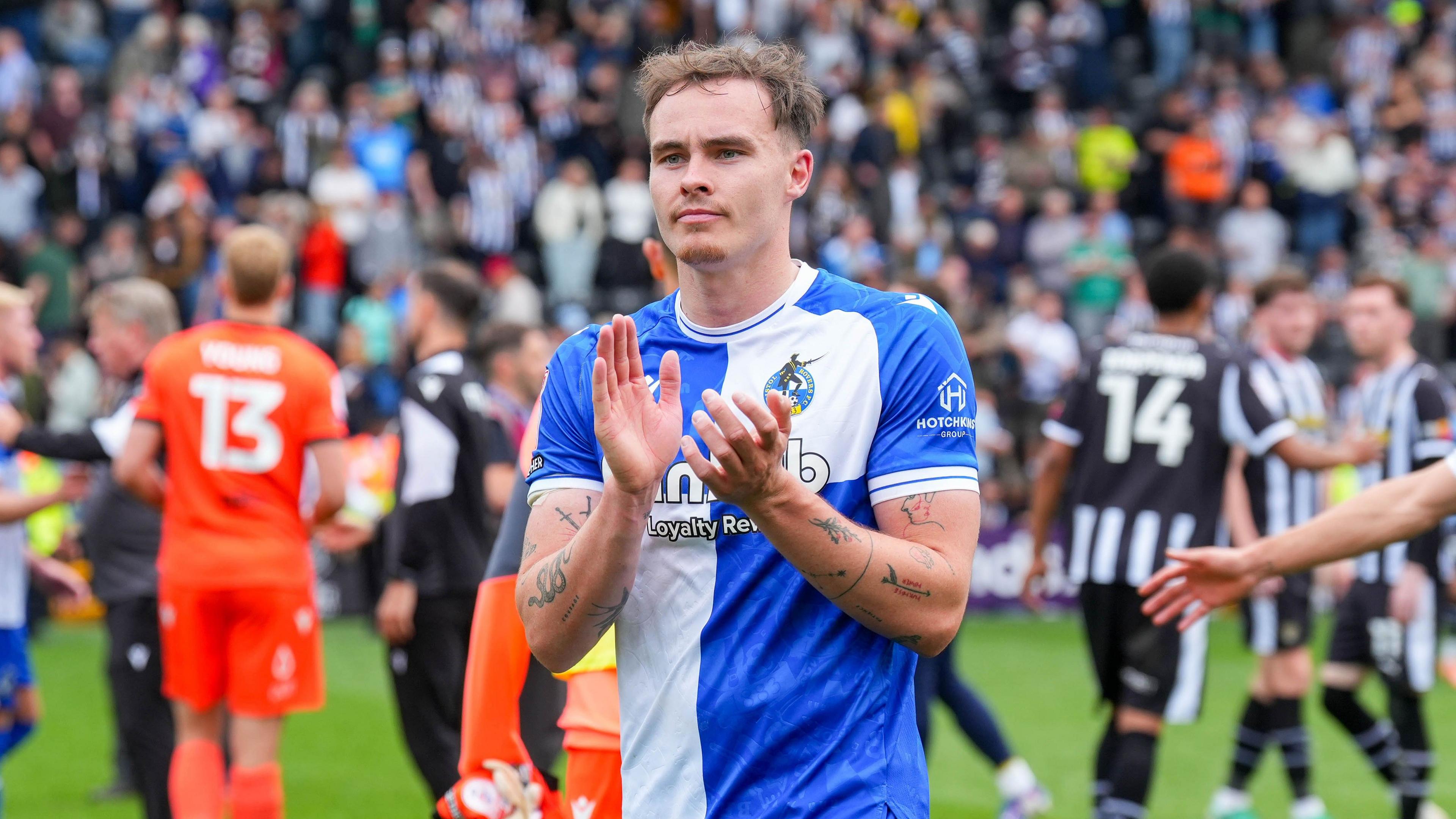 Bristol Rovers defender Kofi Balmer applauding at the end of a EFL football match. Balmer is wearing a blue and white-quartered Bristol Rovers home shirt.