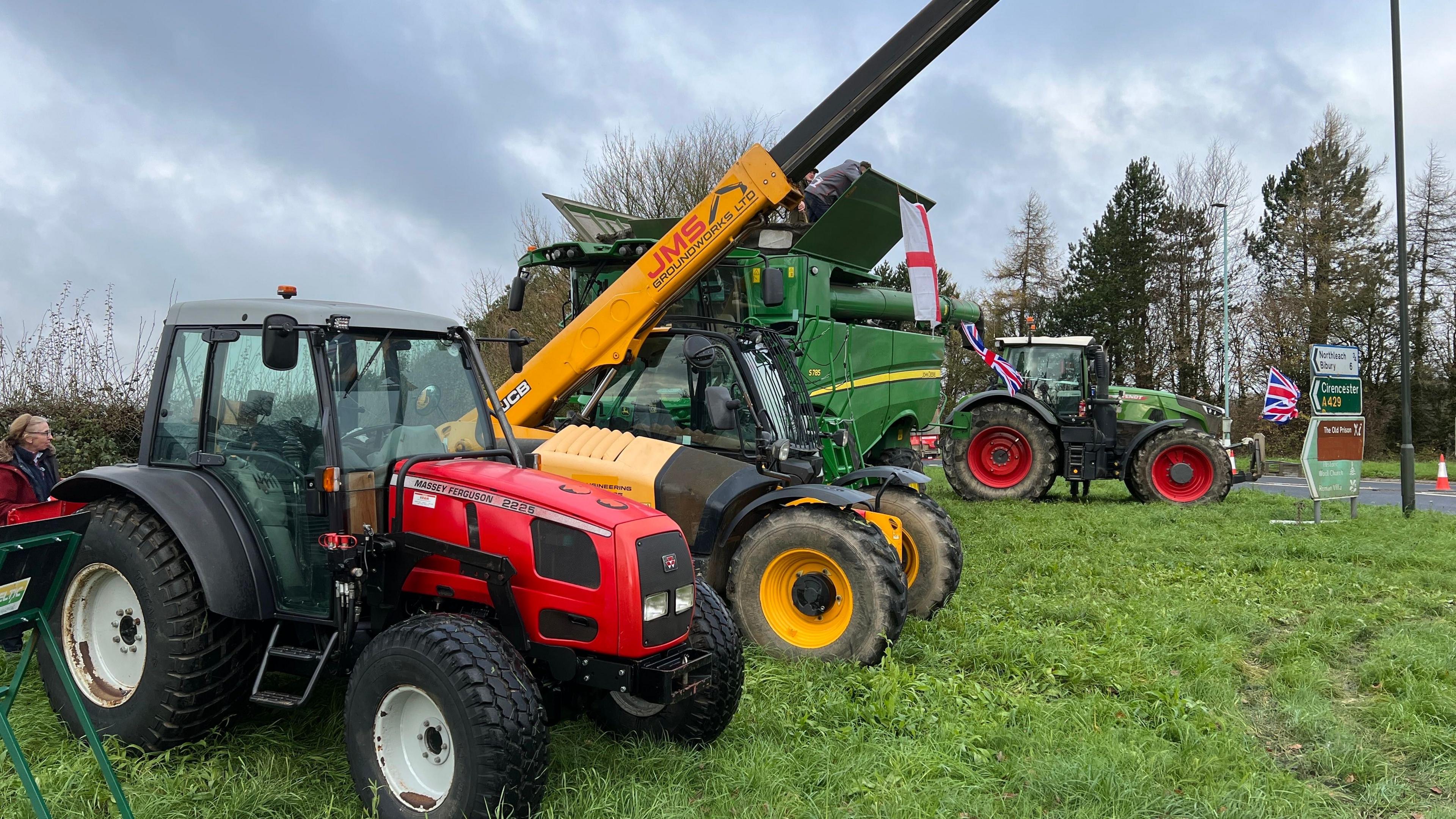 A row of tractors and farming equipment parked on a grassy verge beside a roundabout. There are some St George's and Union Jack flags flying from the vehicles. 