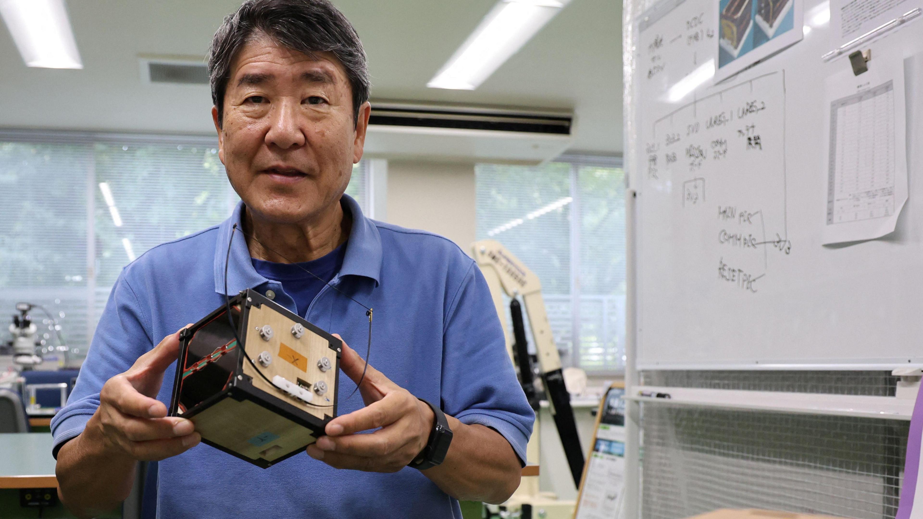 Takao Doi holds LingoSat which is a tiny wooden satellite. It is a small black and timber box with metal fastenings and instruments on it.  He stands in a laboratory in front of a white board with writing and charts on it.