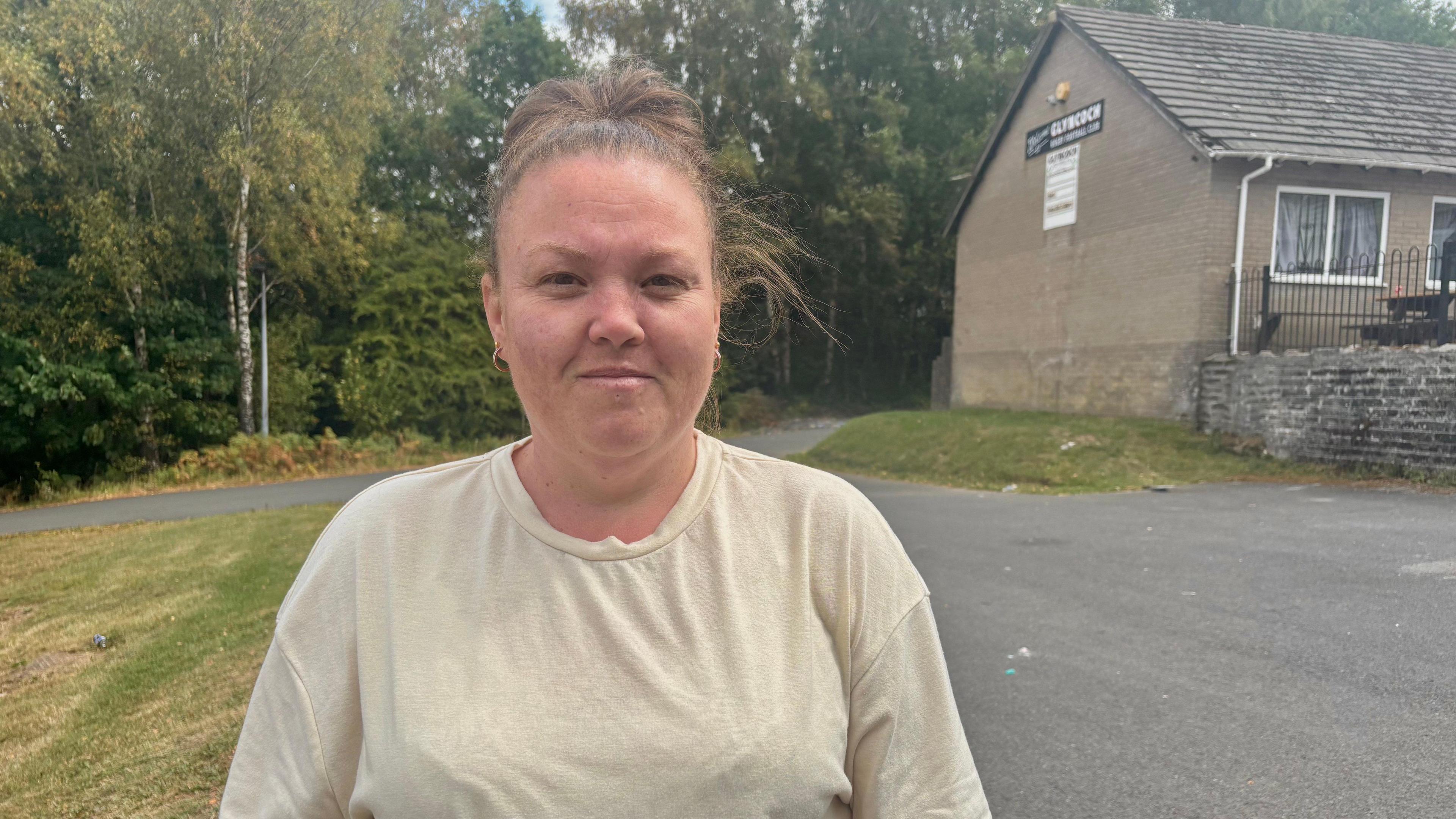 A woman with brown hair tied up is wearing a cream tshirt standing in the car park of a rugby club. Part of the rugby club building can be seen over her left shoulder. There are also some trees in the background.