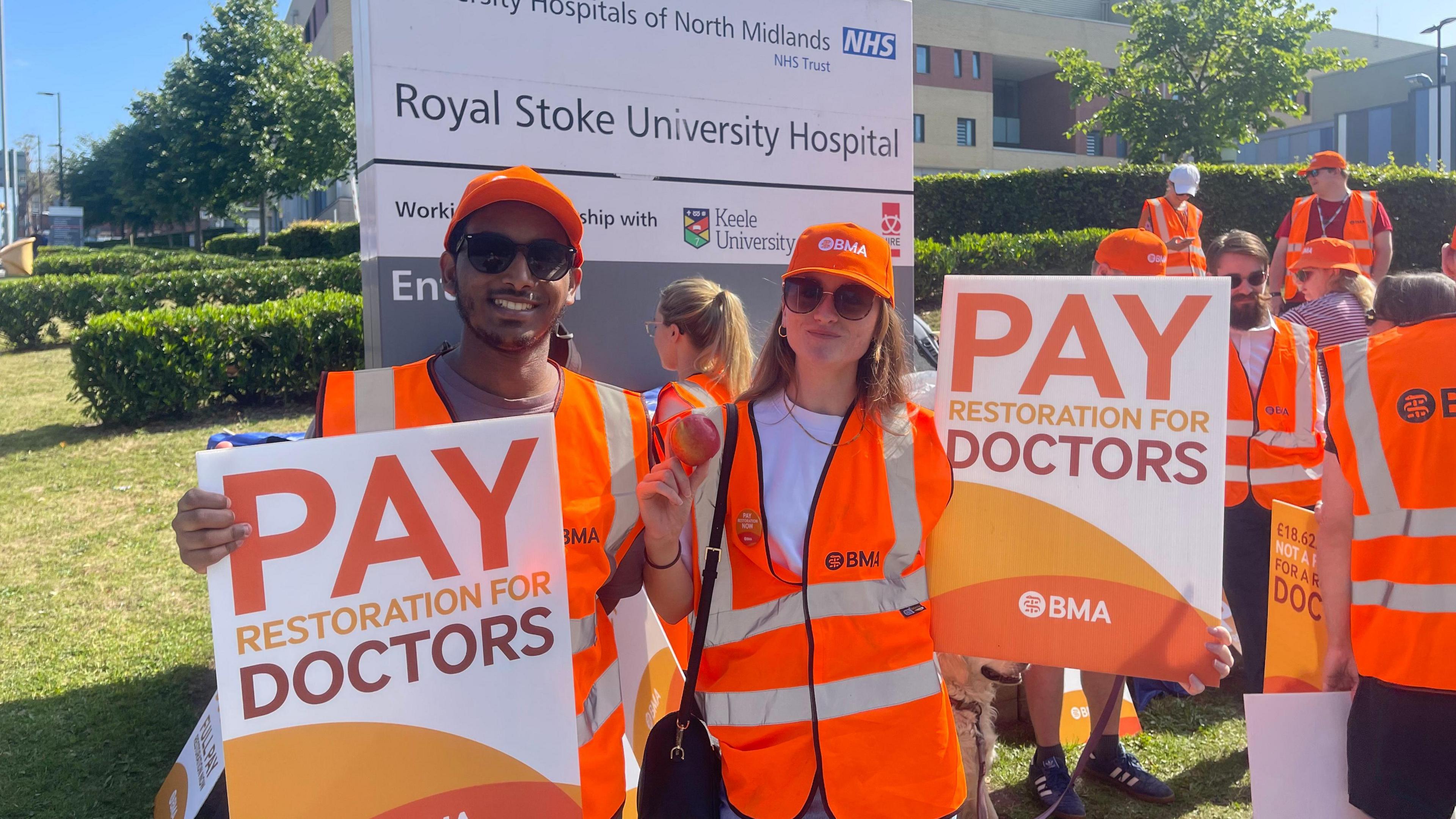 A man and a woman stand outside Royal Stoke Hospital holding placards and signs. The signs read 'Pay Restoration for Doctors'. A large white sign with the words 'Royal Stoke University Hospital' is behind them.