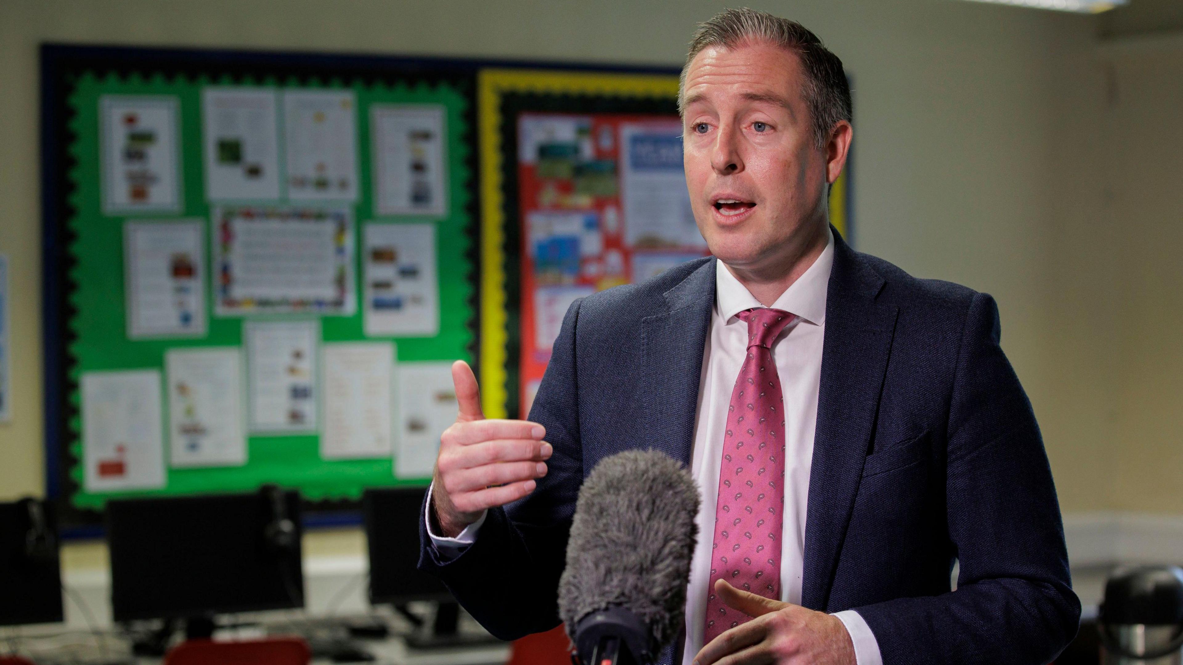 Paul Givan standing in a classroom with display boards behind him. He is wearing a suit and tie and speaking at a microphone, gesturing with one hand out in front of him.